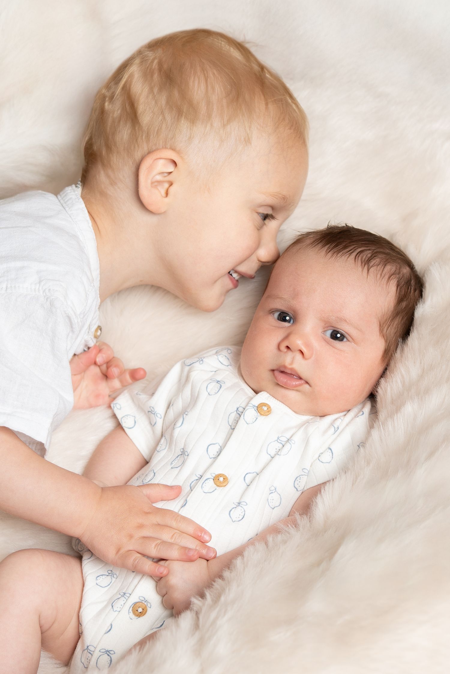 Two babies dressed in white lying on soft white bedding while interacting together in a tender moment.