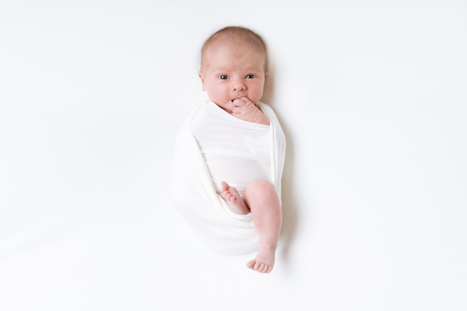 Sweet baby wearing white onesie lies on white background with hand near mouth in peaceful pose.