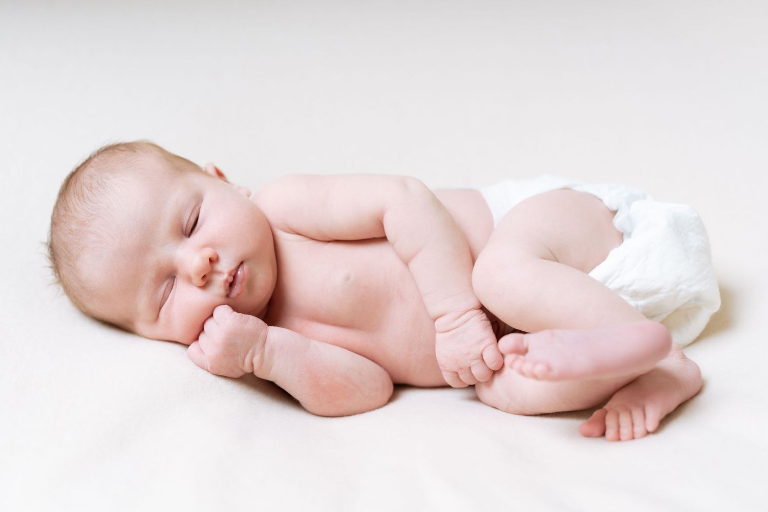 Sleeping newborn wearing white diaper lies on white surface in peaceful curled position.