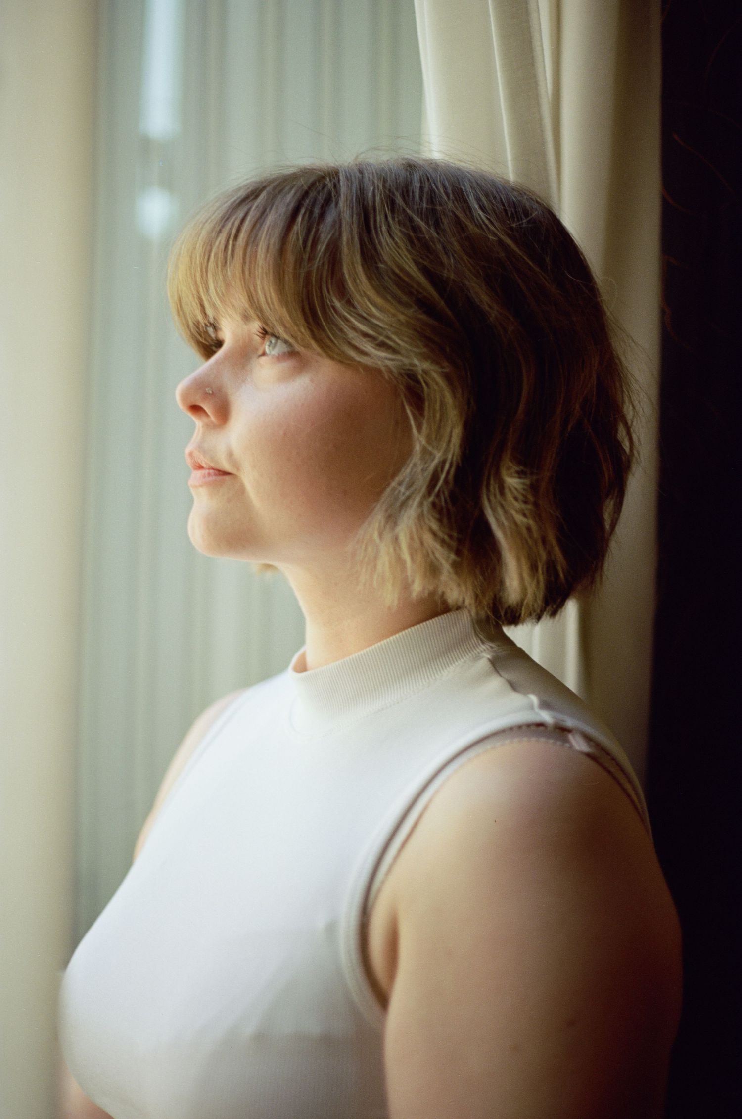 A person gazes thoughtfully out a window in a white sleeveless top, with natural light illuminating their profile.