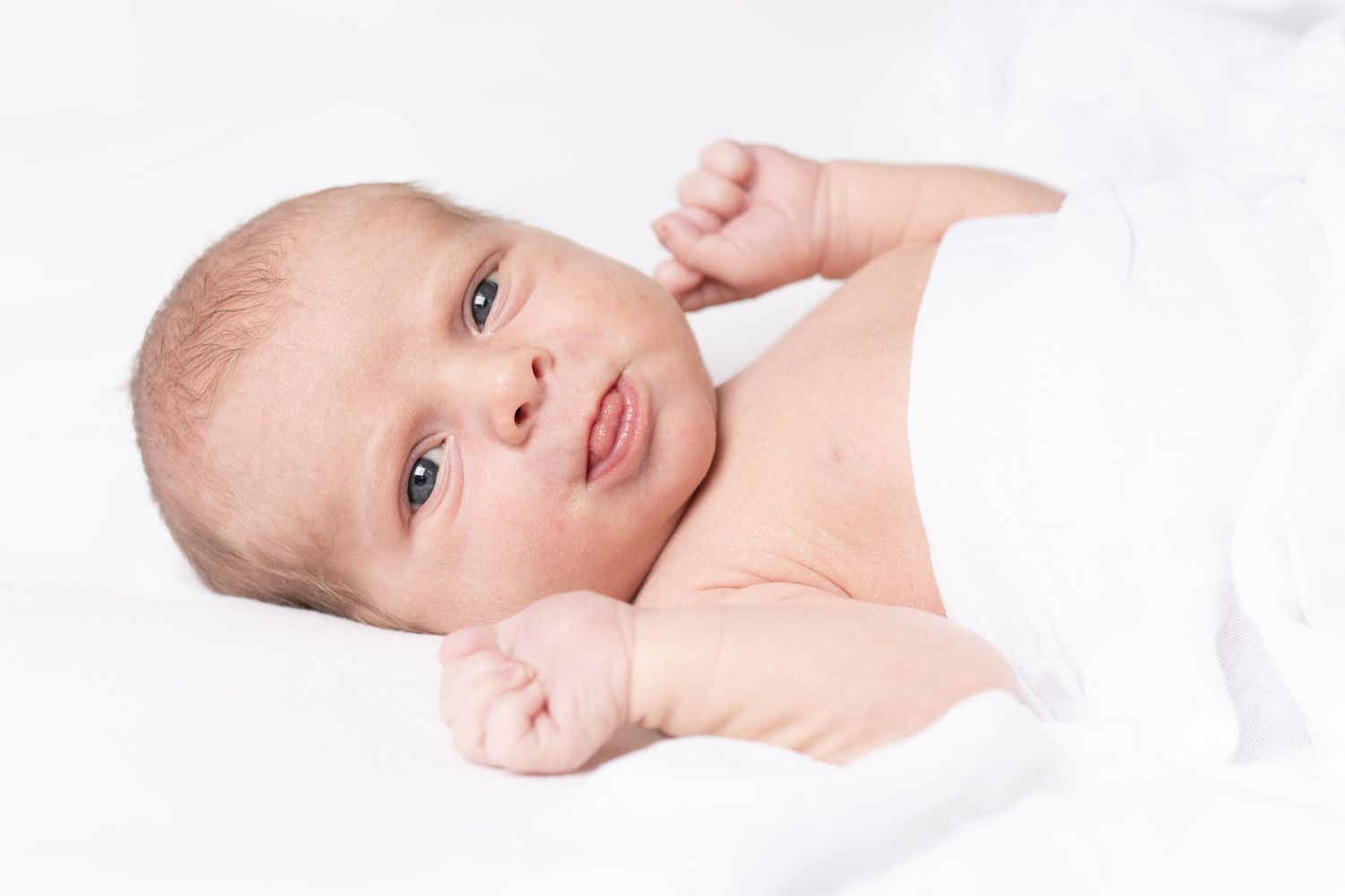 Sweet baby lying on white blanket with wide eyes and pouty lips shows tender expression.