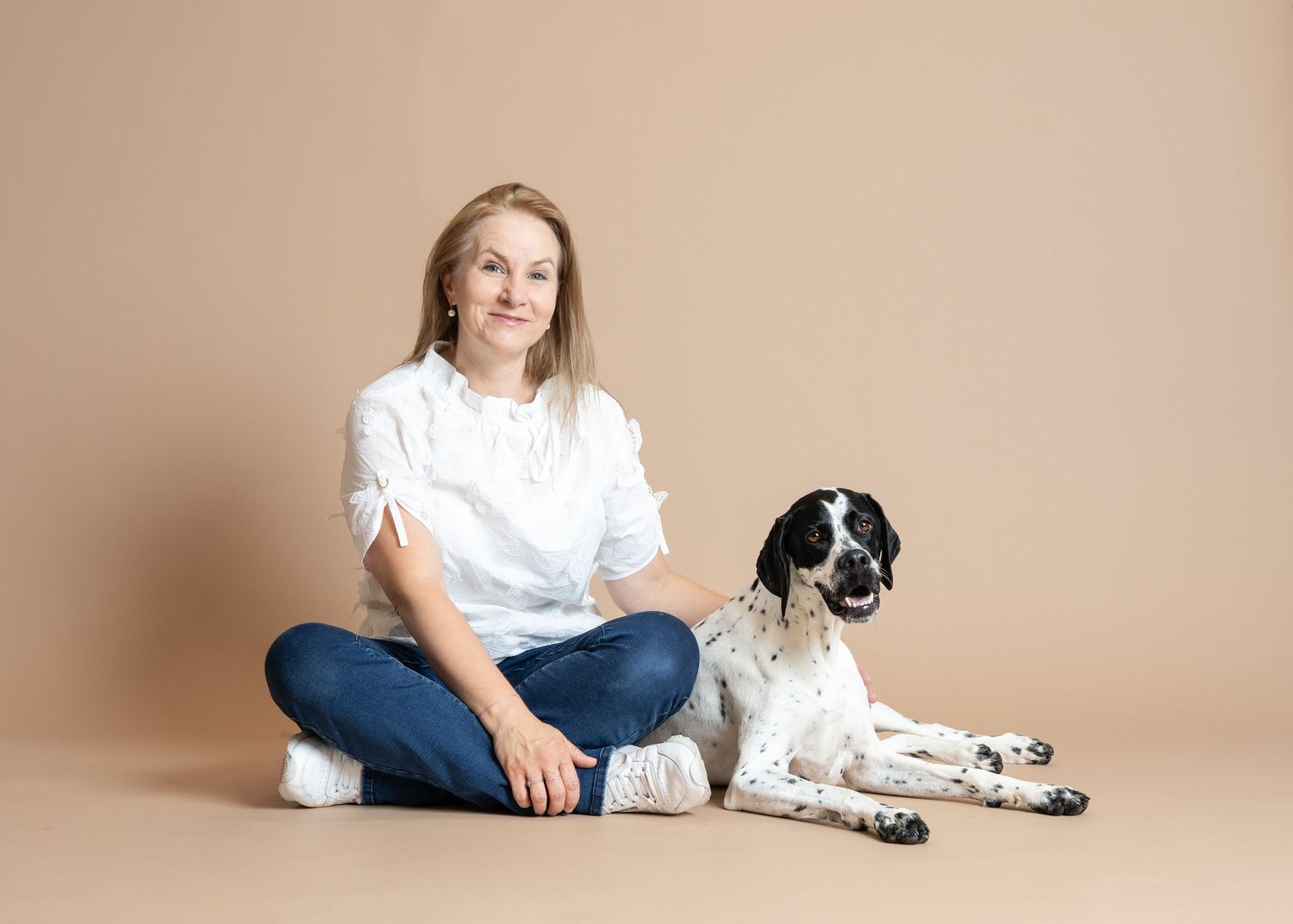 A person in white shirt and jeans sits cross-legged on beige backdrop next to black and white spotted English Setter dog.