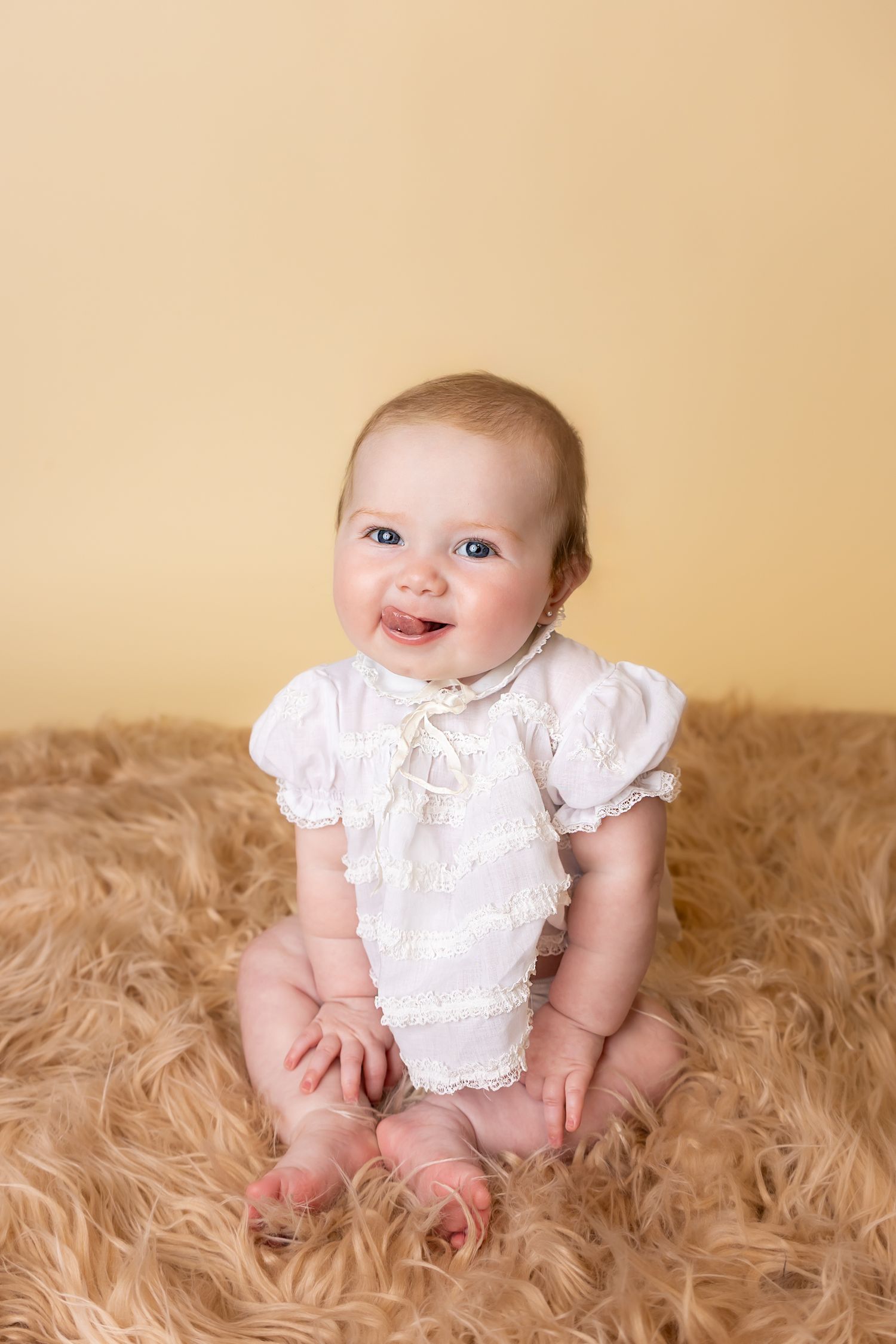A baby in a vintage white lace dress sits on a beige carpet and sticks her tongue out during a mini session in Pittsburg Ks and Joplin MO.