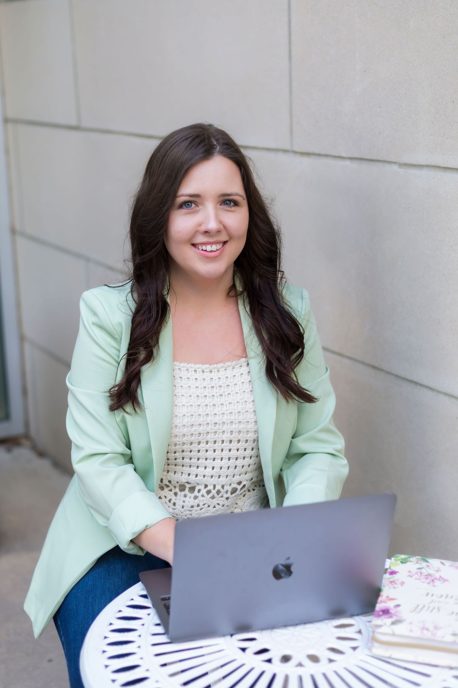 Professional working outdoors with laptop on white table wearing mint green blazer and white crochet top.
