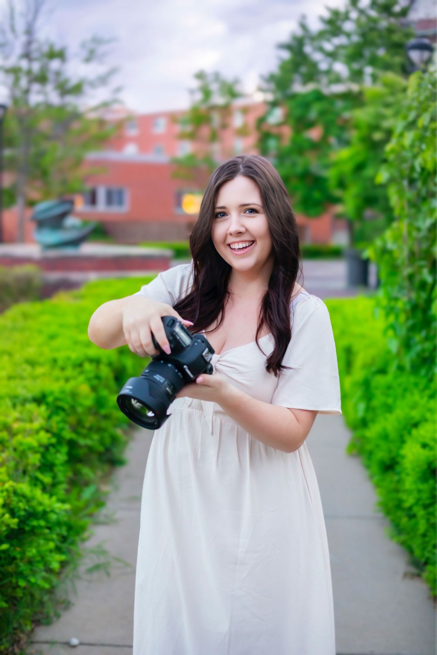 Professional family photographer in Pittsburg KS and Joplin MO, wearing a white dress and posing outdoors with a camera.