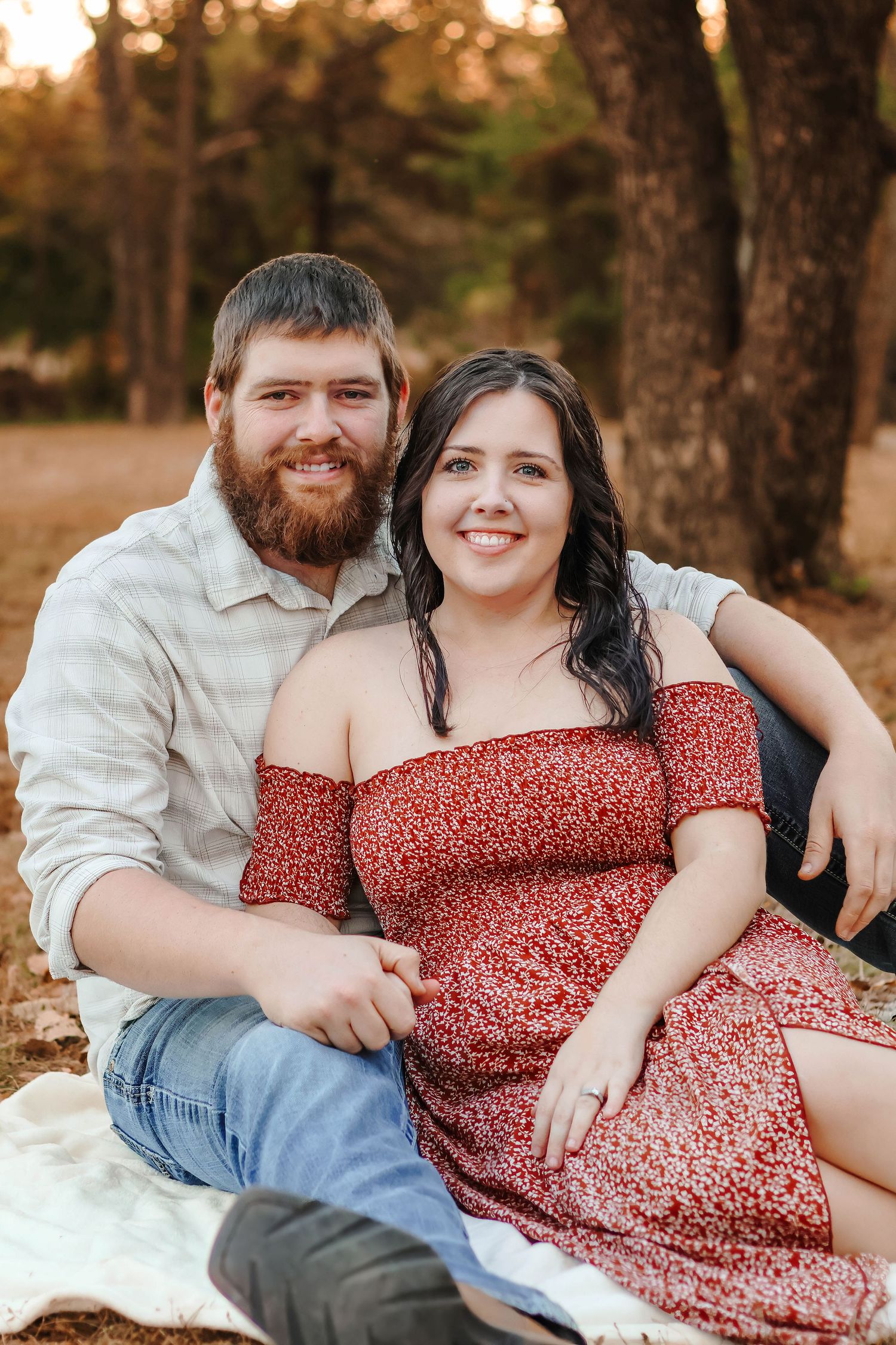 Couple sitting on blanket outdoors during golden hour in casual attire with trees in background.