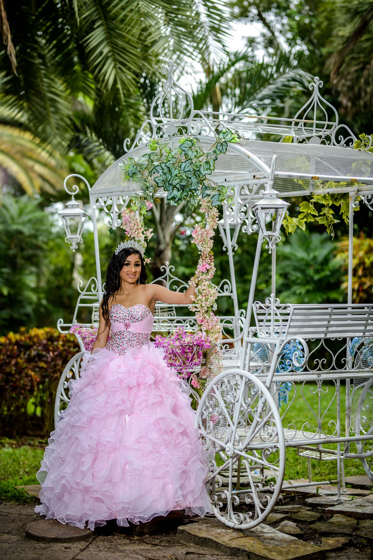 Ornate white carriage gazebo with floral decorations in a lush tropical garden setting features pink ballgown and palm trees.
