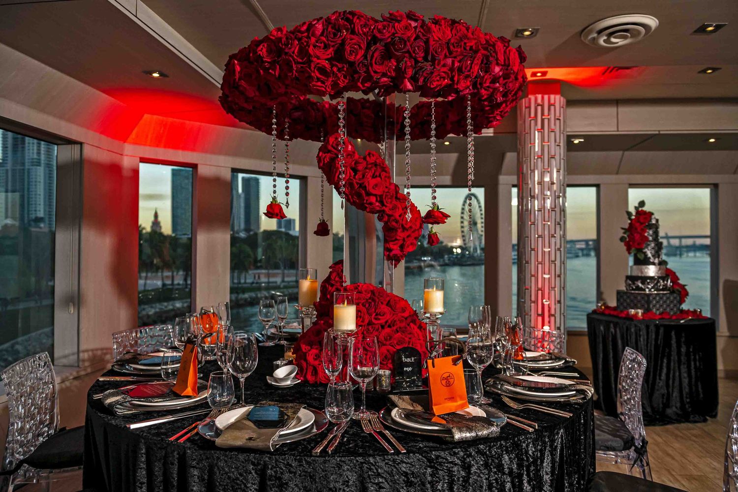 Formal table setting with suspended red rose arrangement and black linens in waterfront venue.