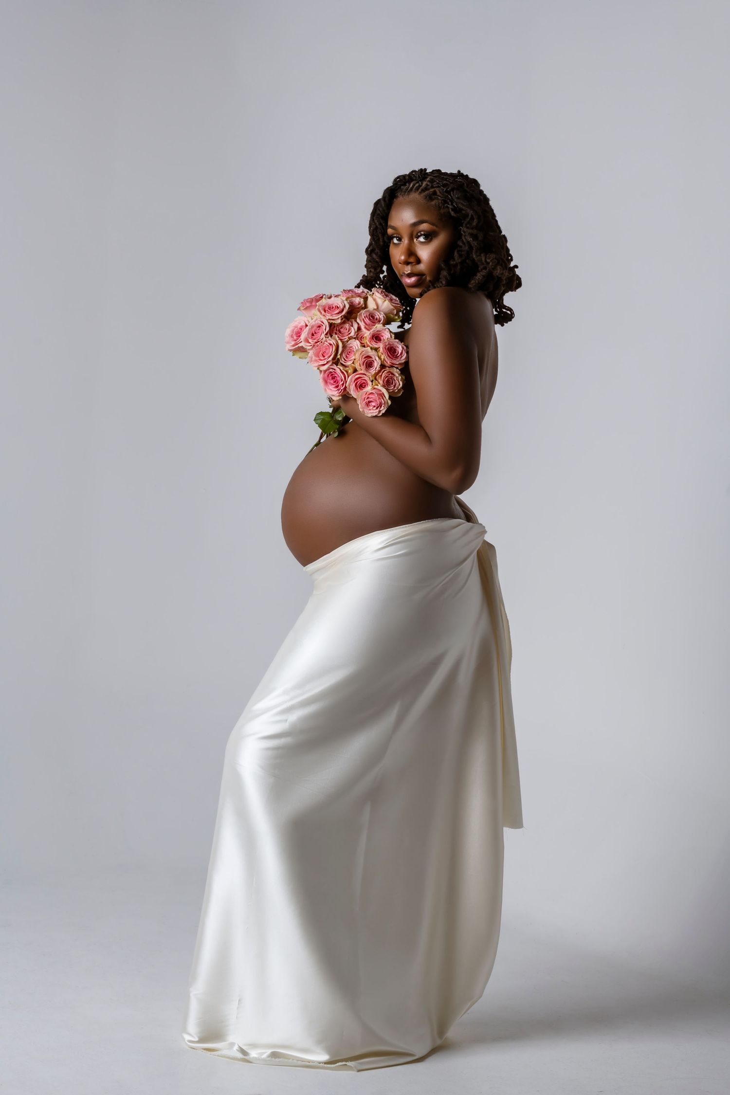 Artistic maternity photoshoot featuring white fabric draping and pink roses against grey backdrop.