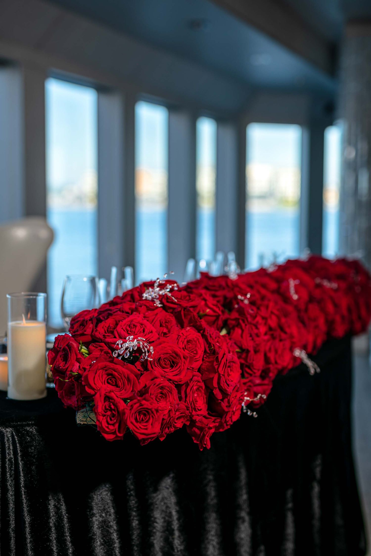 Long table adorned with deep red roses and glowing candles against a waterfront window backdrop on the Seafair Mega Yacht in Miami.