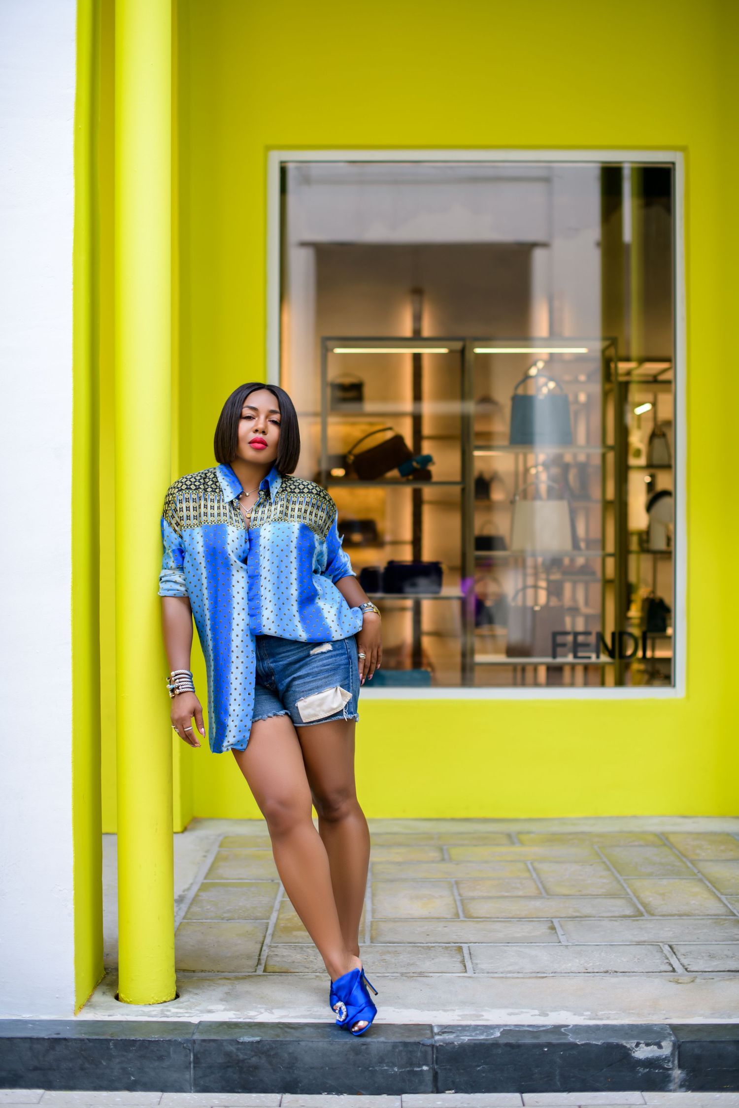 A model wearing a blue striped shirt and denim shorts poses outside a bright yellow storefront with a display window.