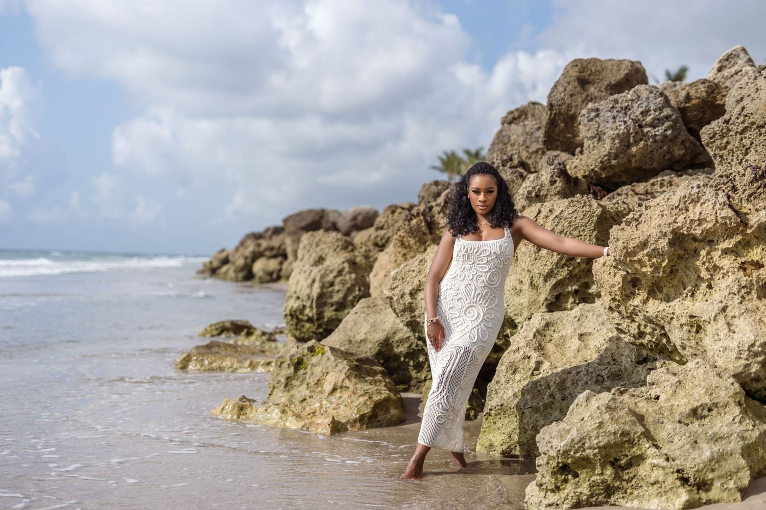 Person in white dress poses elegantly against limestone cliffs on a sunny beach.