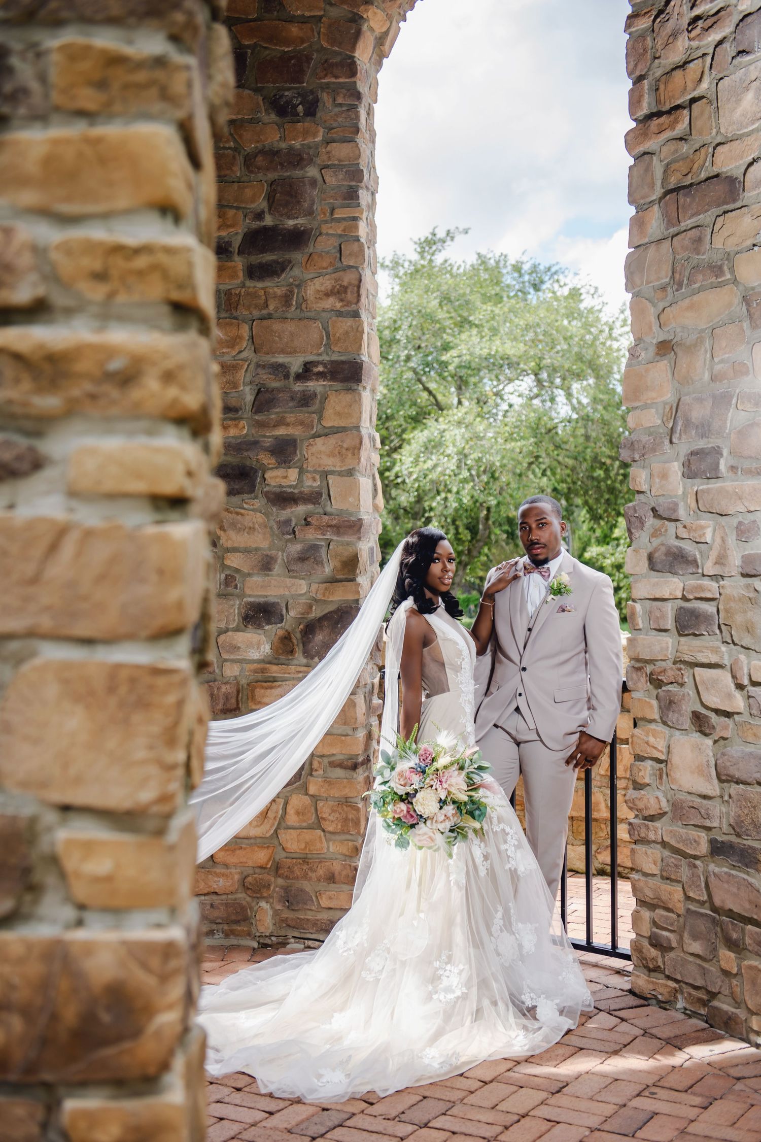 A newly married couple poses between stone pillars outdoors, with the bride wearing a flowing white gown and long veil.