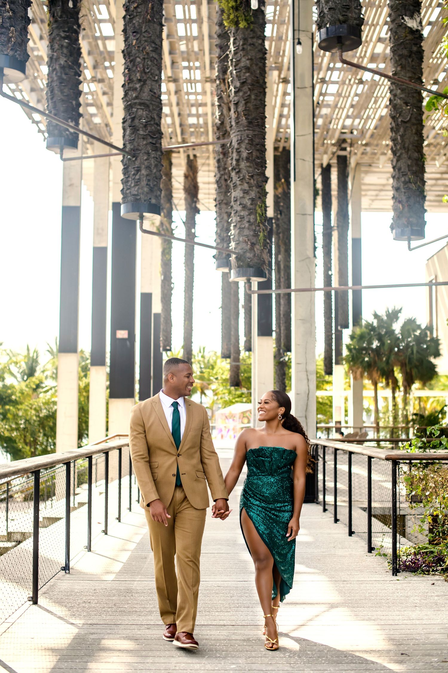 Elegant couple walking hand in hand beneath a row of palm trees in an outdoor walkway.