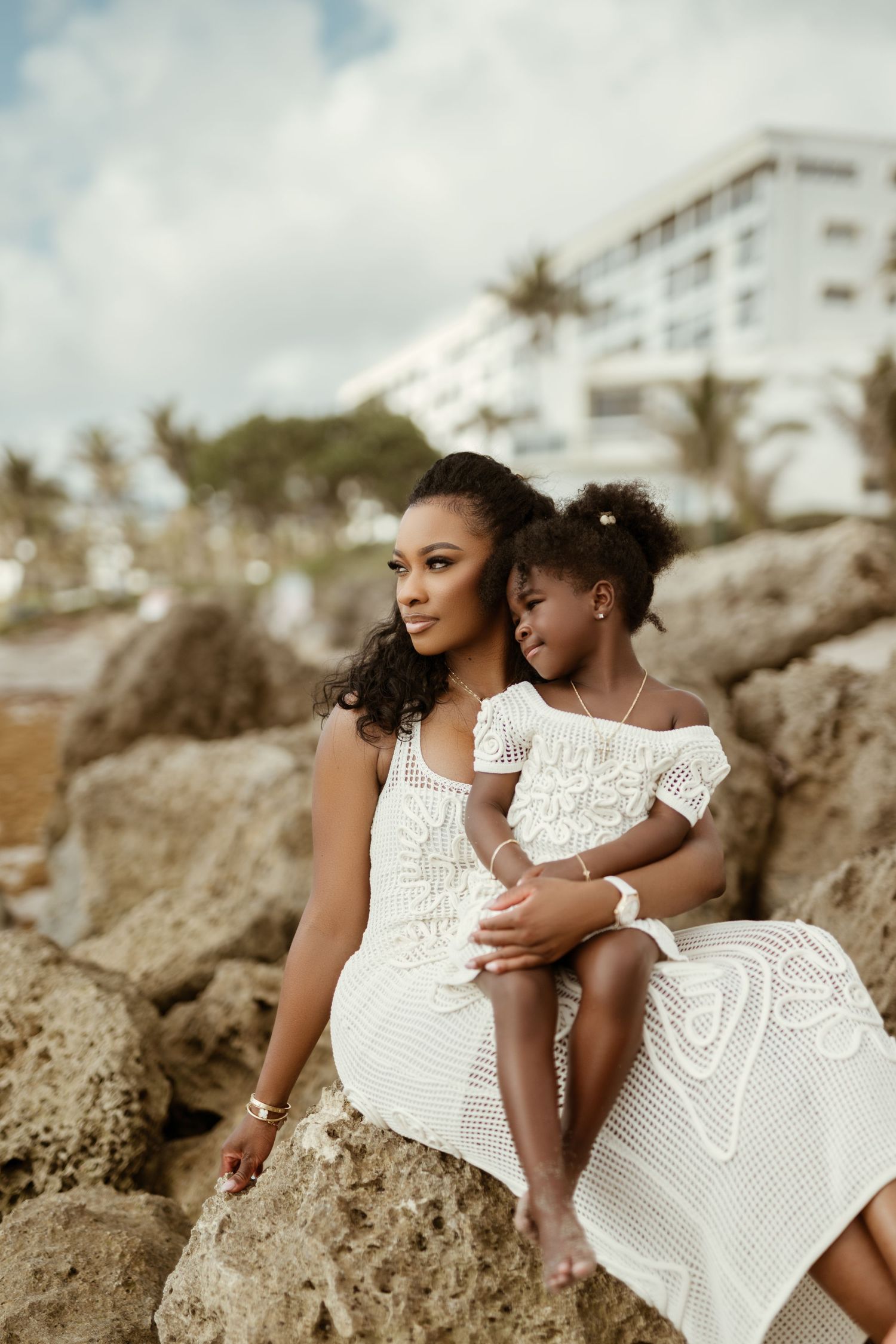 Mother and daughter in white dresses sitting together on coastal rocks by the ocean.