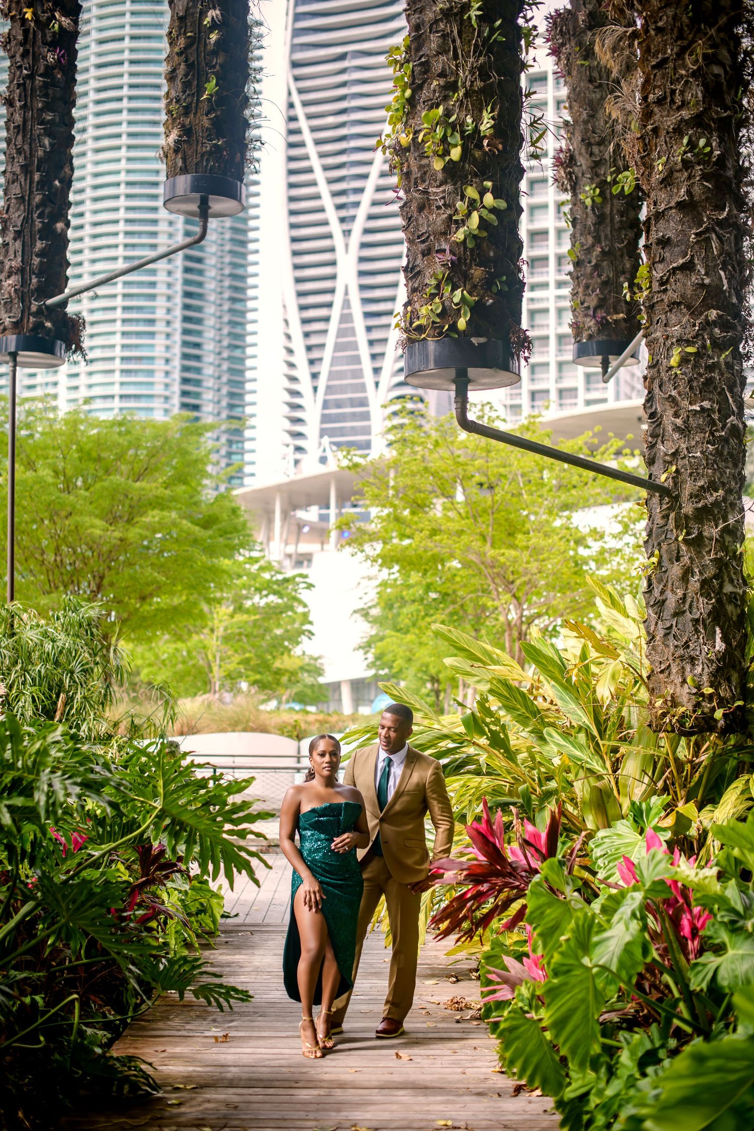 A couple in formal wear strolls through a lush tropical garden path with skyscrapers in the background.