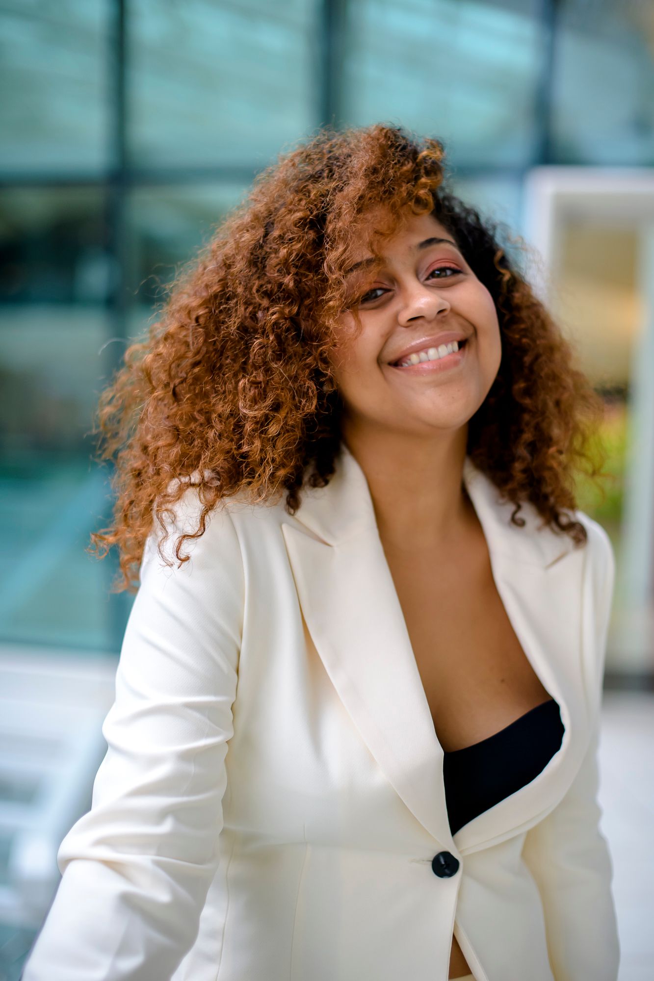 Professional woman in white blazer with natural curly hair smiling outdoors.