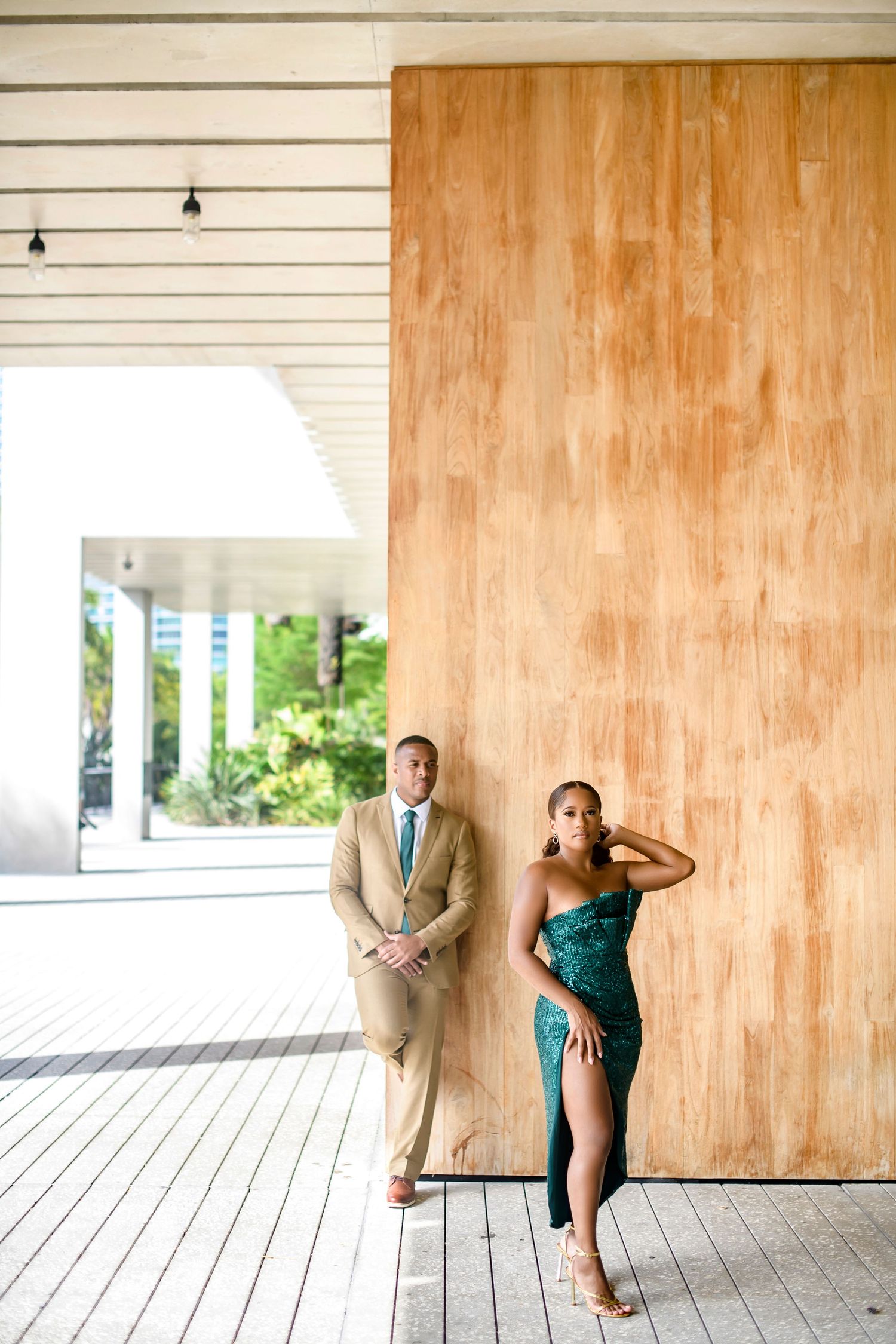 Stylish couple in formal attire posing against wooden wall in minimalist setting.