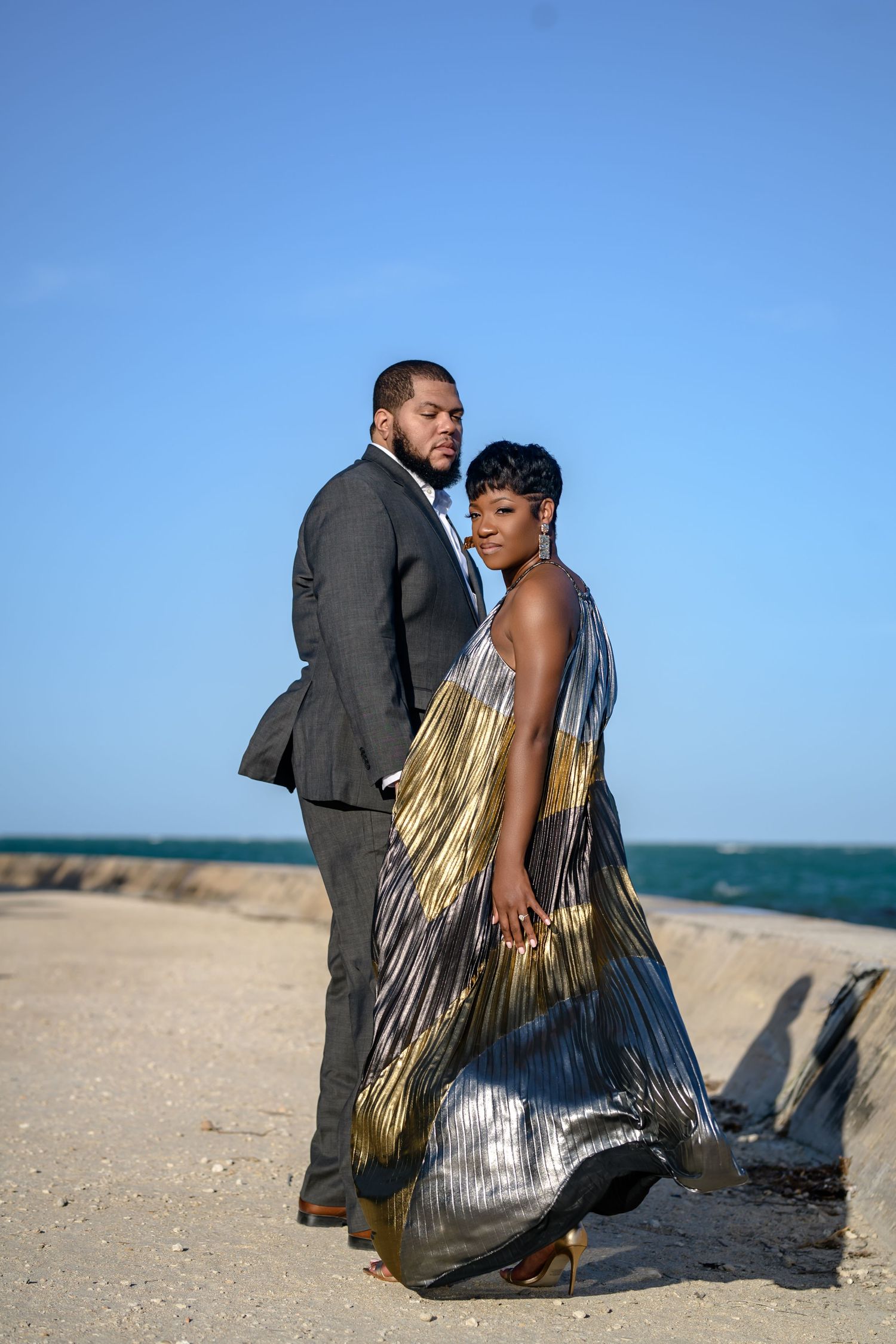 A couple in formal attire poses romantically on a beach, with the woman wearing a flowing metallic dress against blue sky.