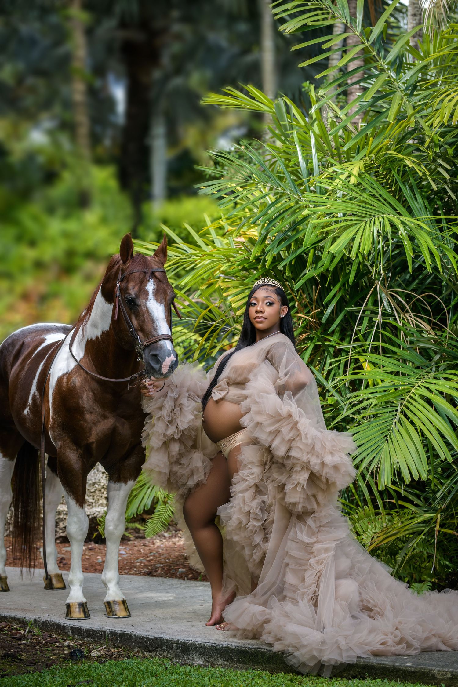 A brown and white horse stands next to a flowing beige tulle gown against a lush tropical palm background.