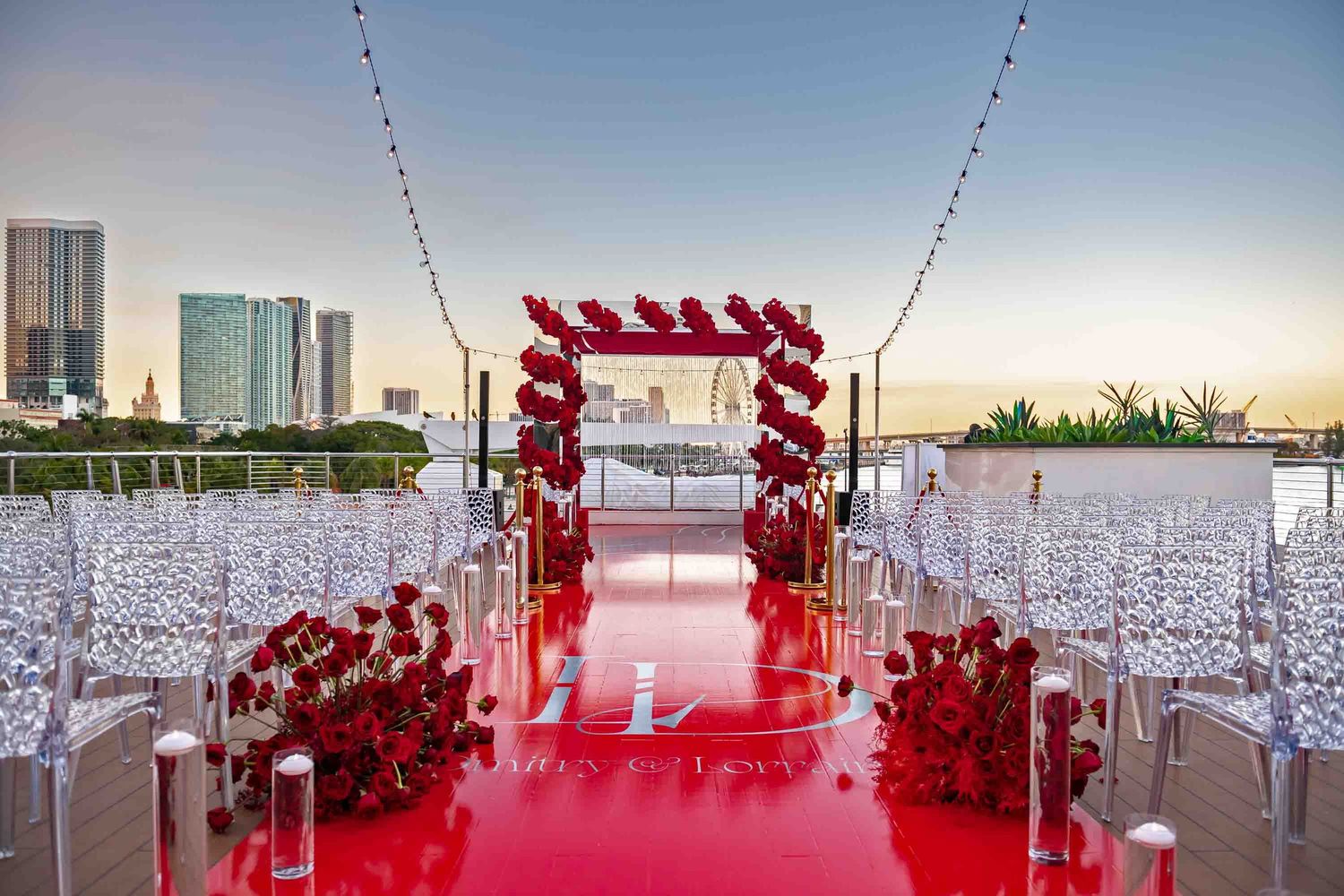 Wedding aisle decorated with red roses and arch against cityscape at sunset.