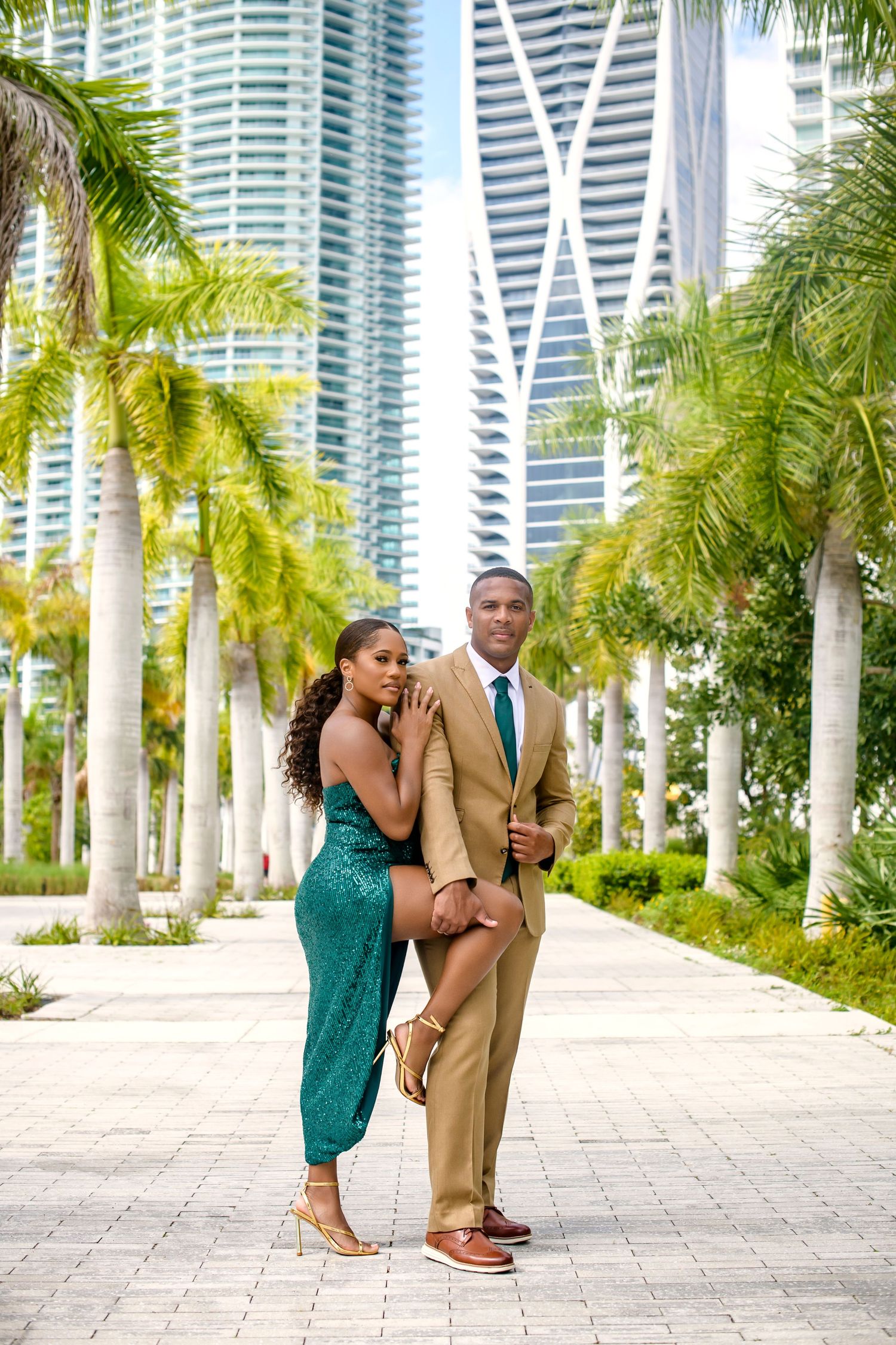 Glamorous couple in formal wear posing along palm-lined path with modern skyscrapers in background.