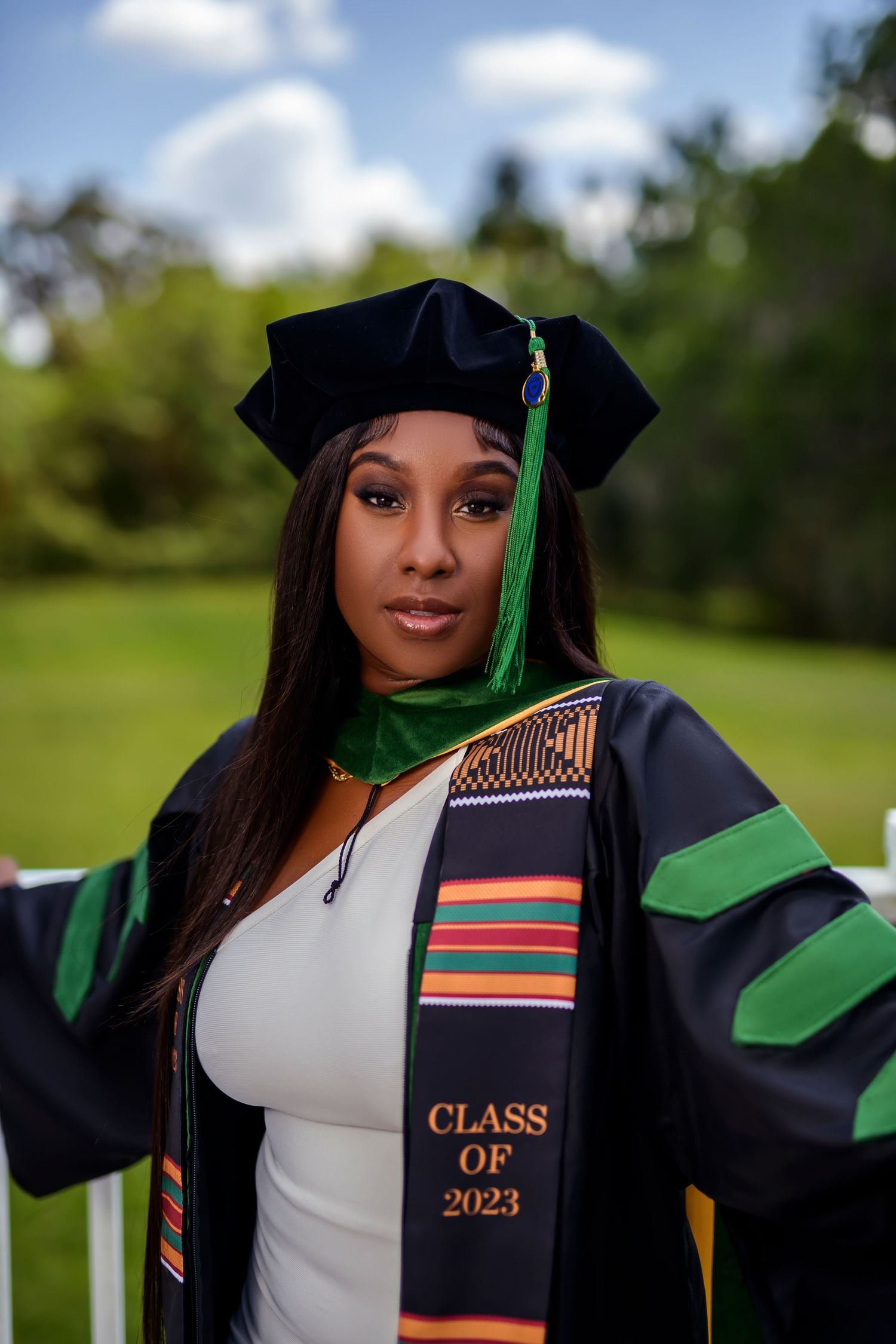 Graduate wearing academic regalia with Class of 2023 sash poses outdoors in front of greenery.