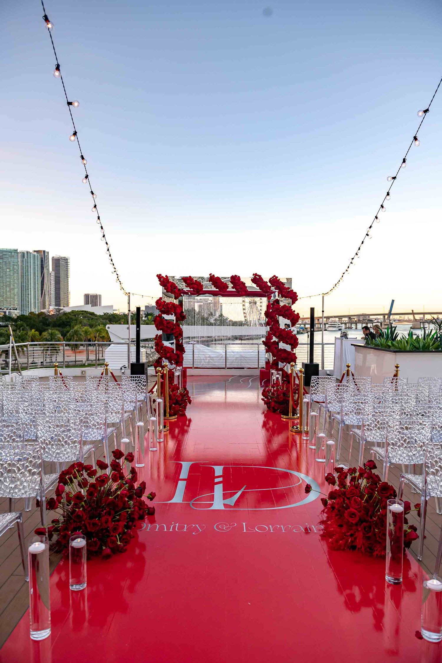 Red carpet aisle with balloon arch and white chairs for rooftop wedding ceremony against city skyline on the Seafair Mega Yacht in Miami.