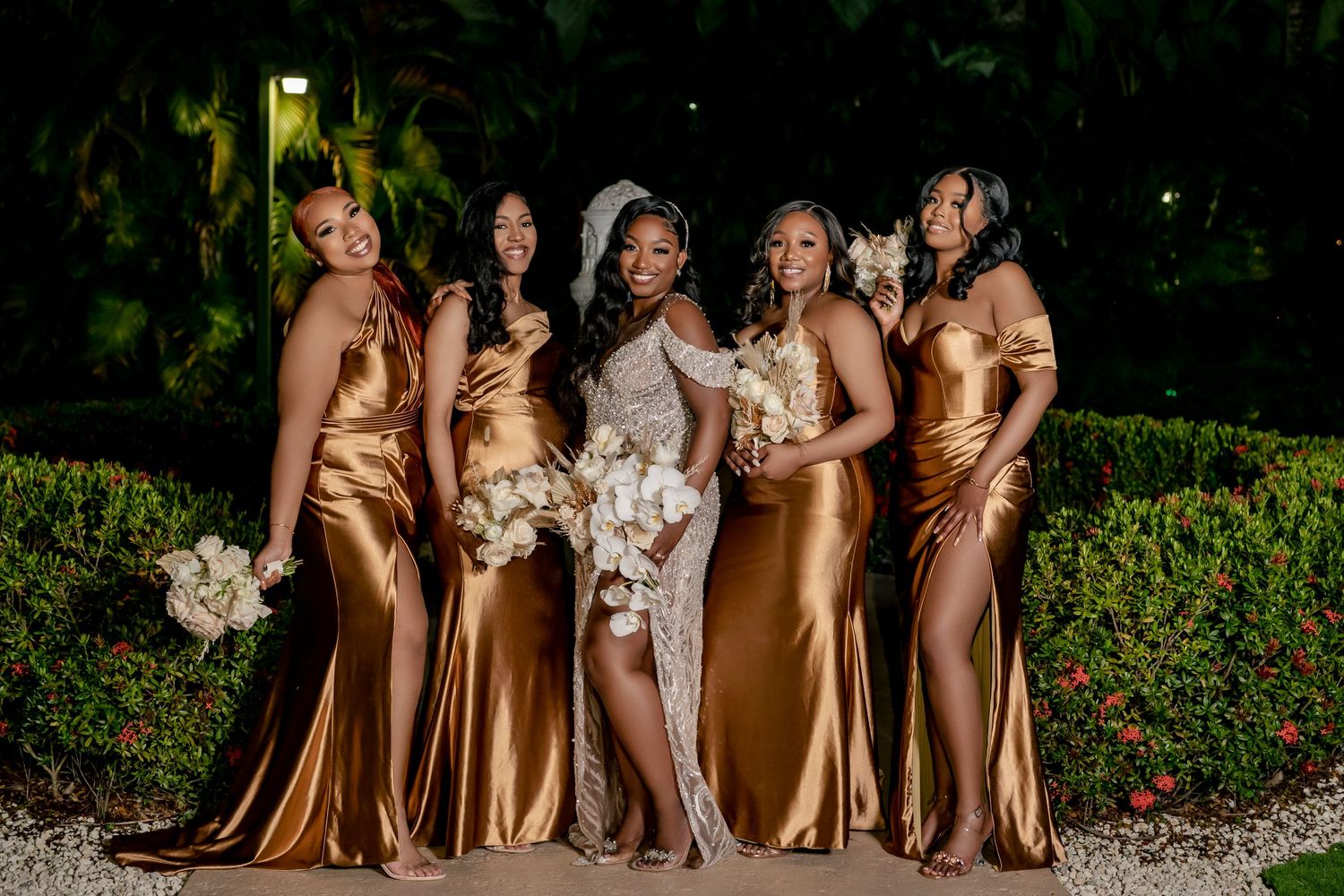Wedding party in gold bridesmaid dresses and white bridal gown pose outdoors at evening event.