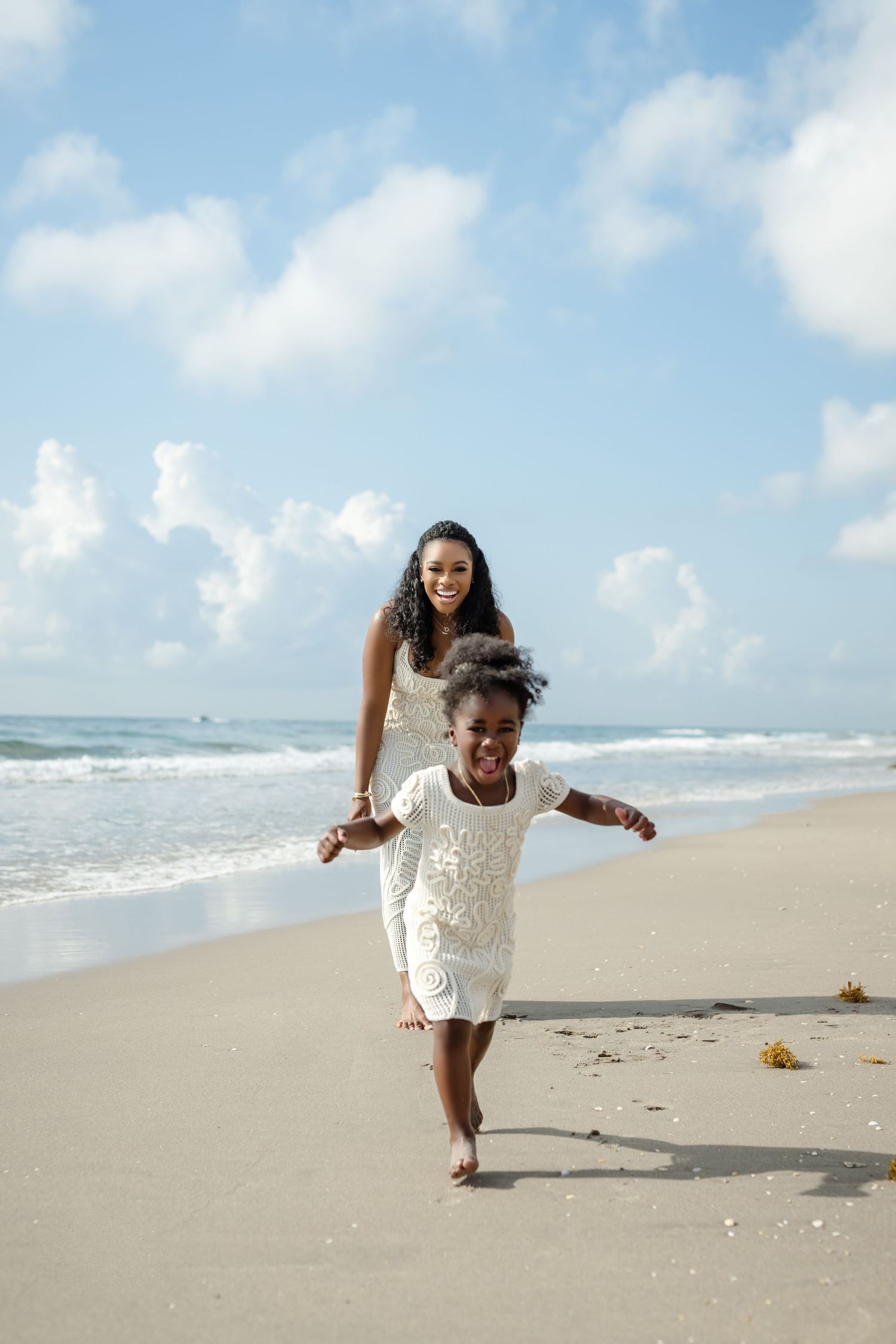 Two people in white dresses run playfully along a sunny tropical beach.