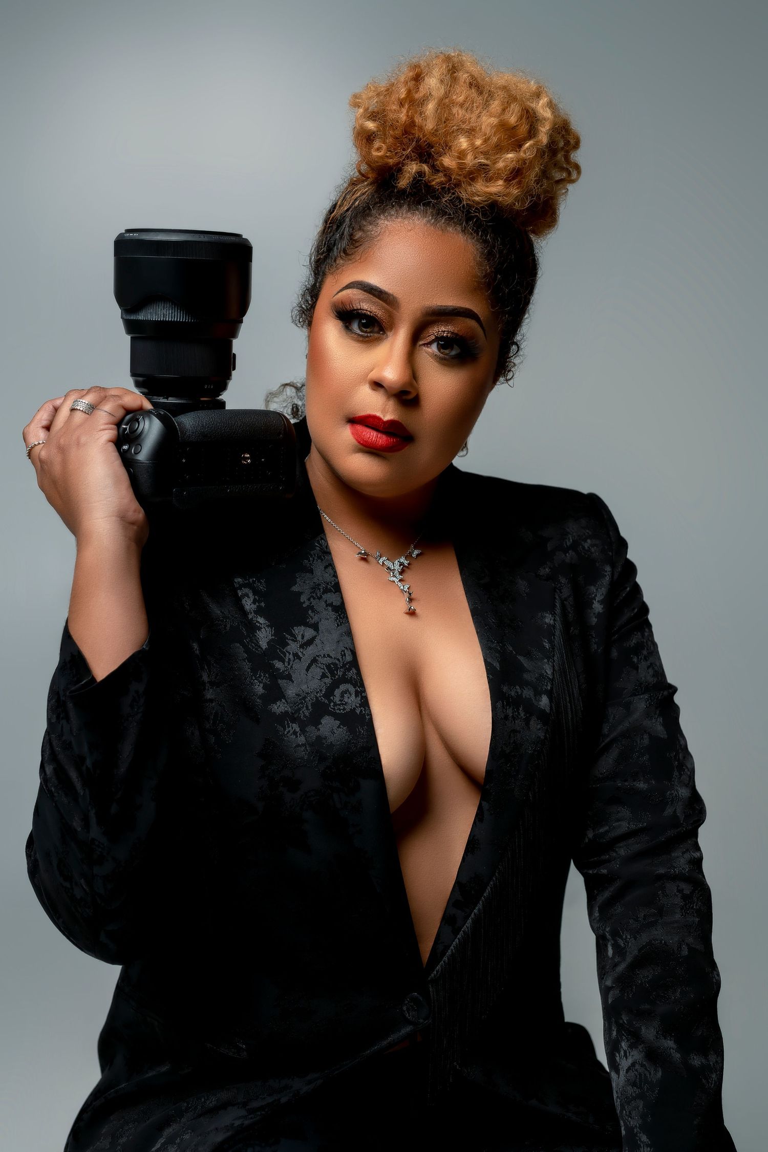 Portrait of professional photographer in black blazer holding camera against grey background.