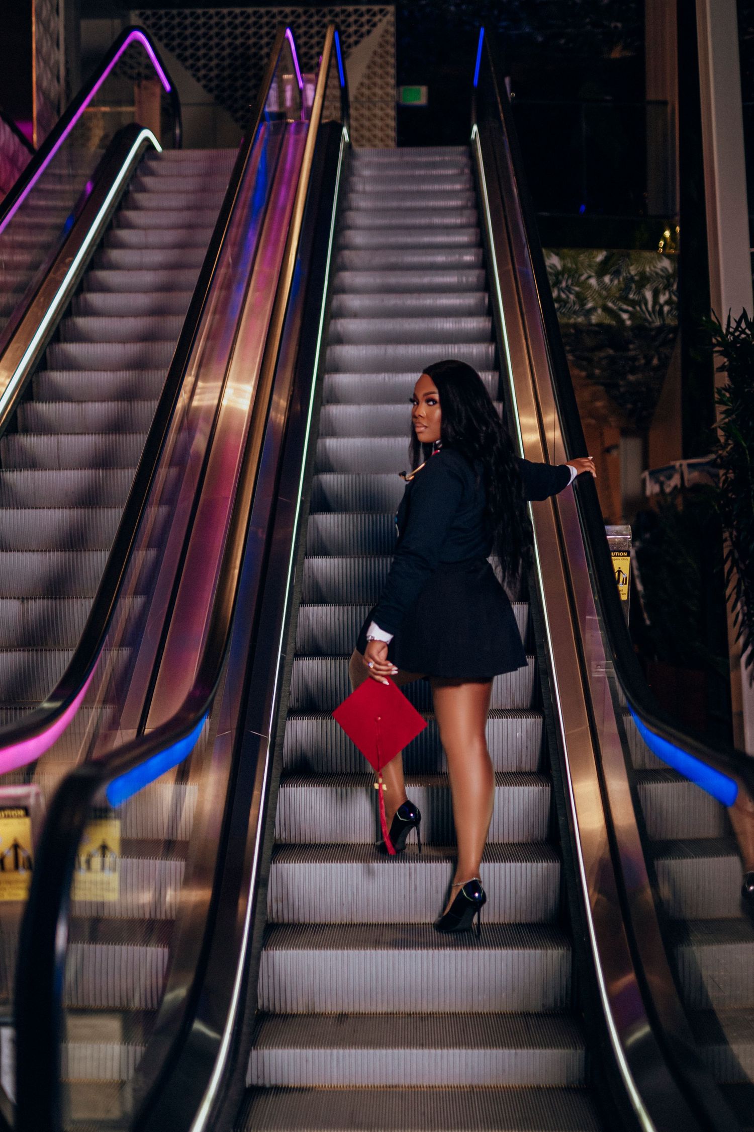 Stylish woman in black on illuminated escalator at night with neon colored handrails and red purse.