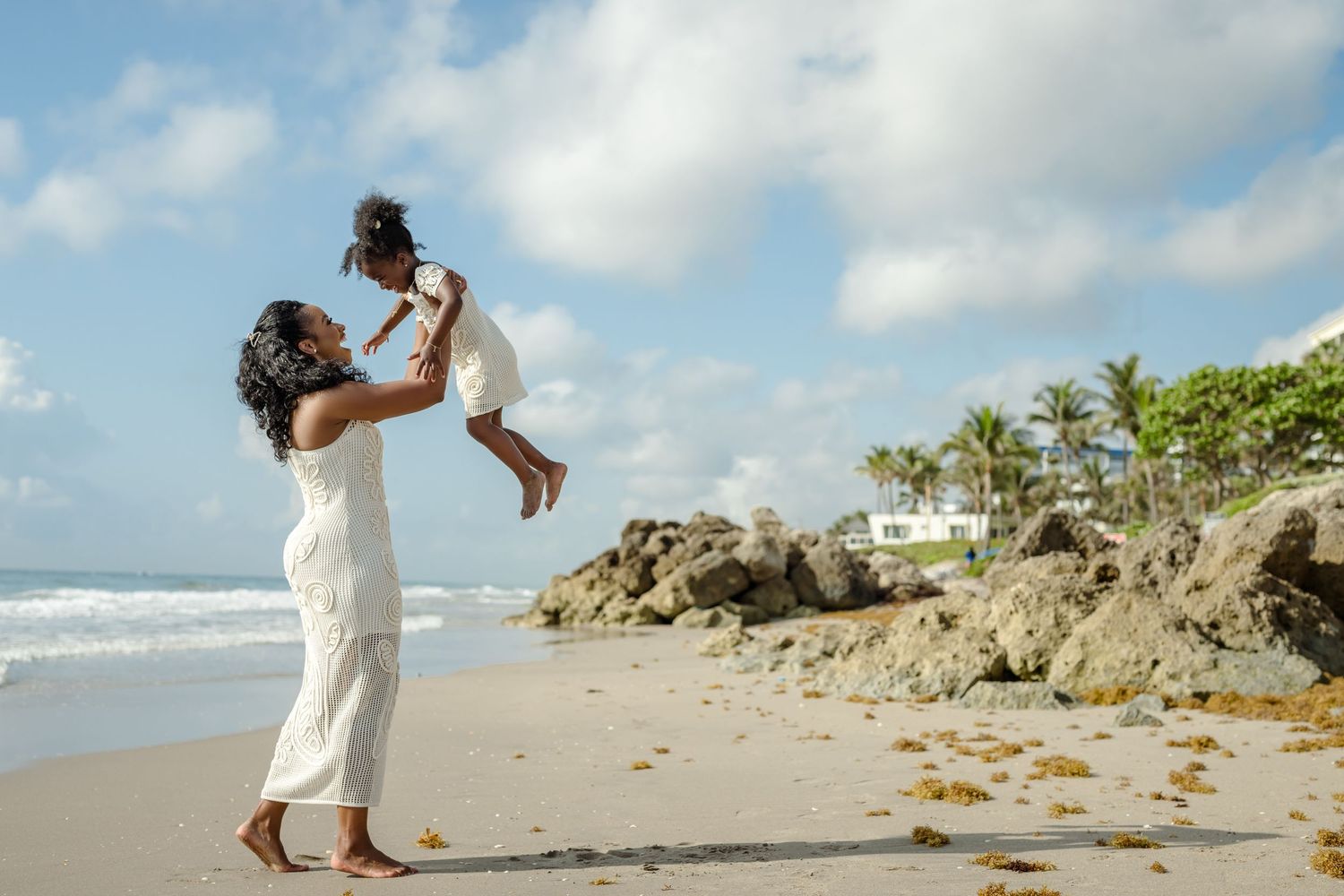 Two people in white dresses play joyfully together on a scenic beach with rocky cliffs and palm trees.