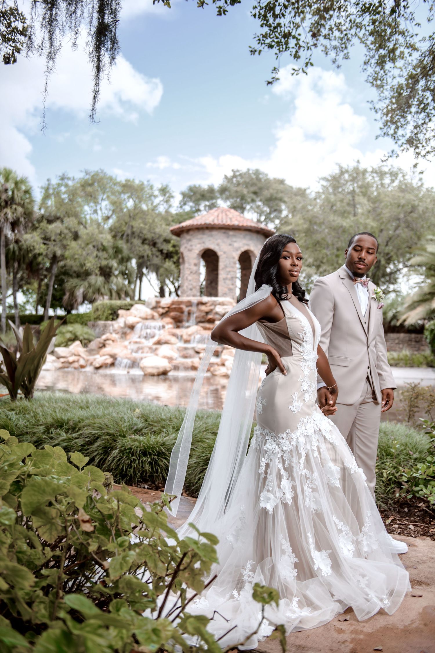 Couple in wedding attire pose in garden setting with stone gazebo.