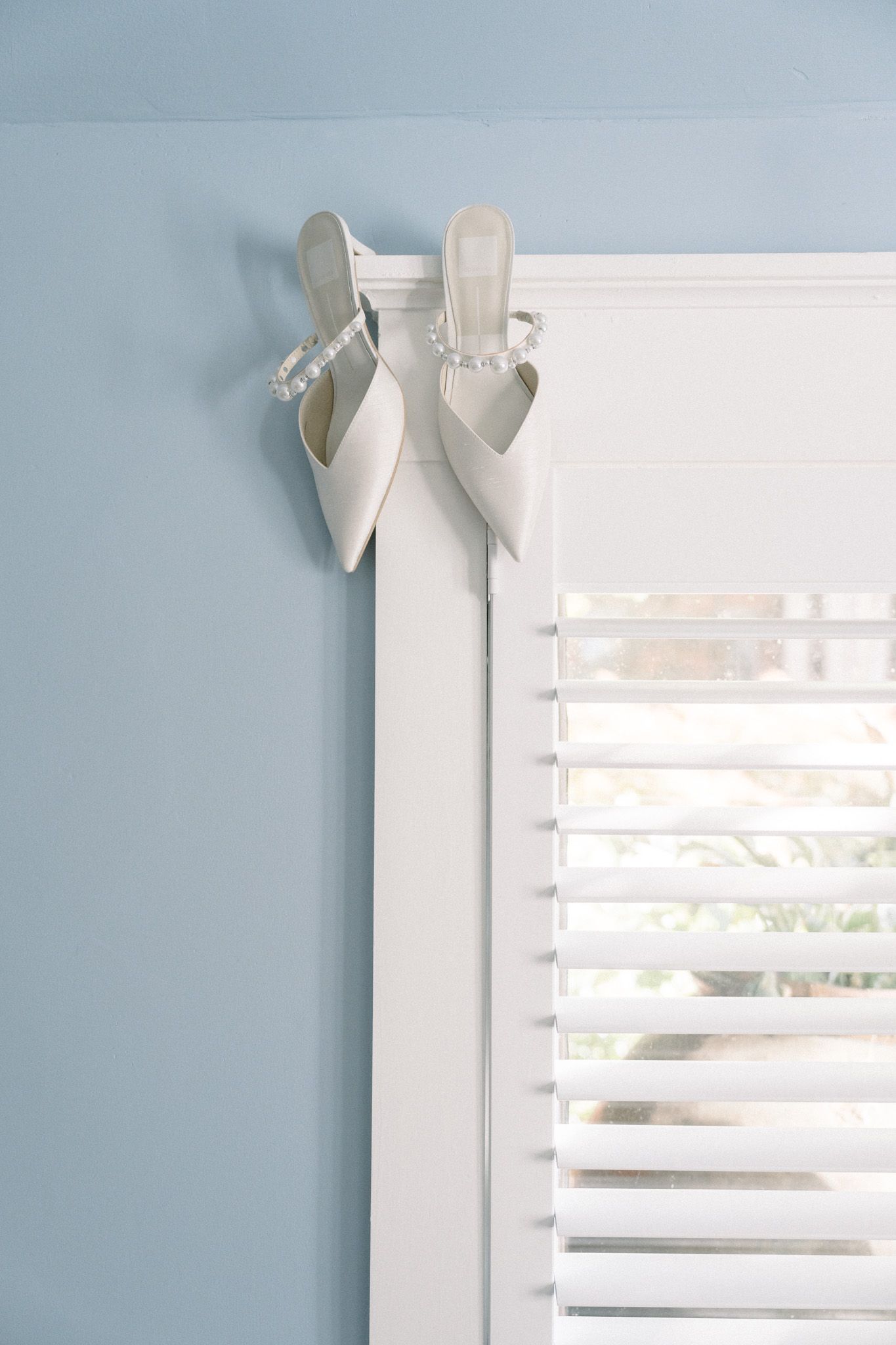 White high heel wedding shoes hanging decoratively on a window frame with white blinds against a pale blue wall.