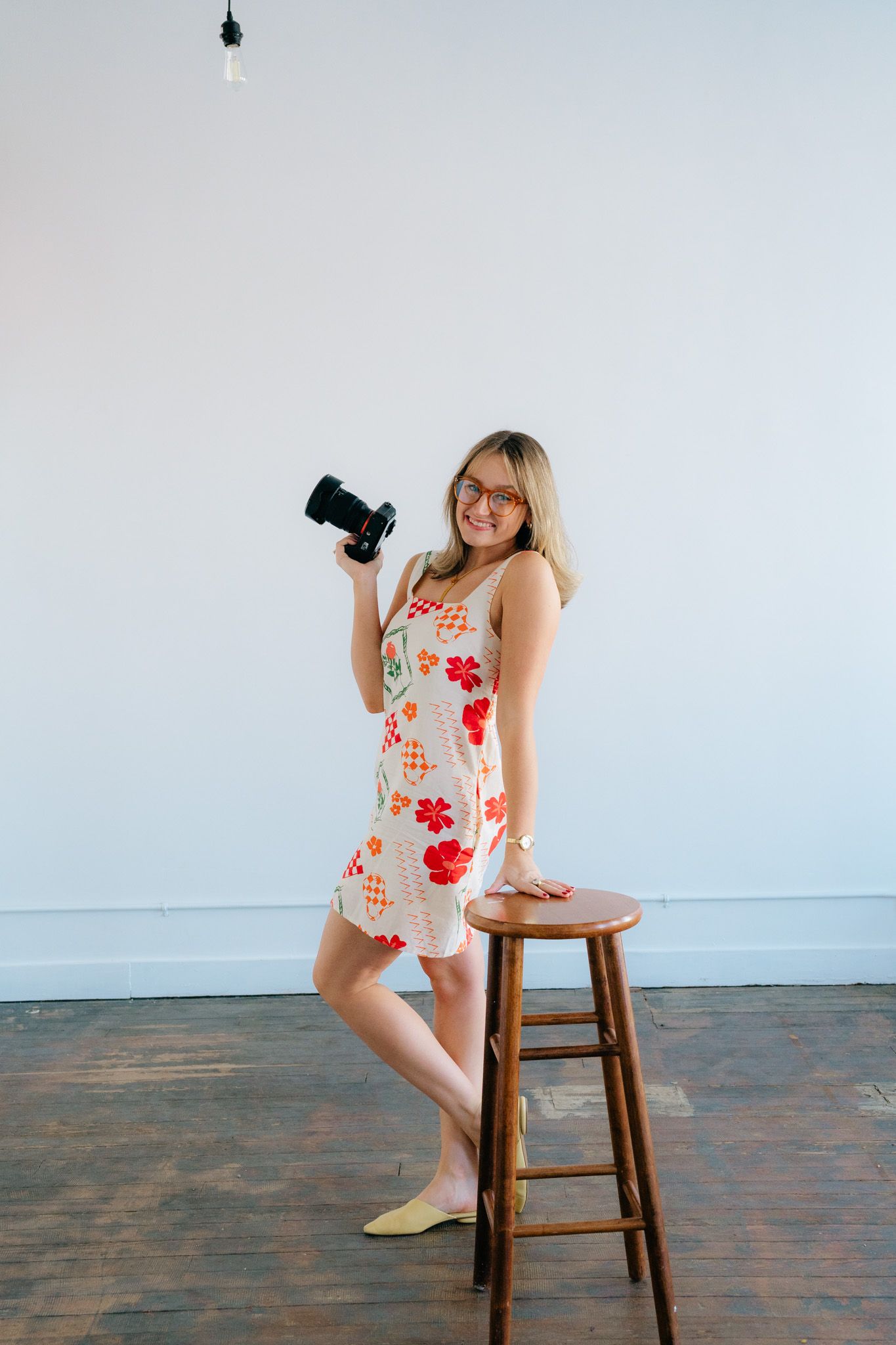 Professional photographer in a floral summer dress poses with camera next to wooden stool in minimalist studio setting.