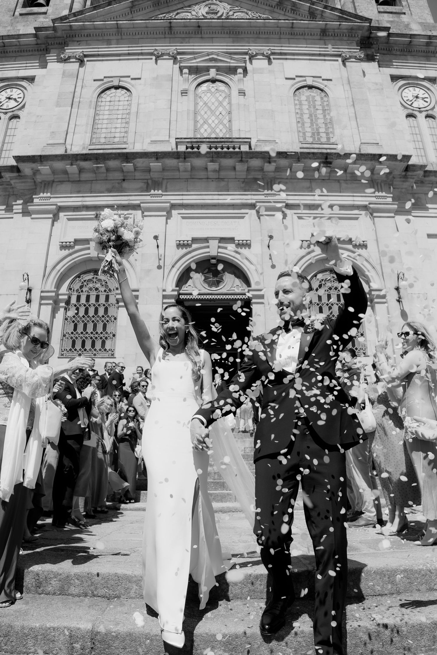 A wedding celebration outside a grand church as guests throw confetti over a couple exiting through ornate doors.