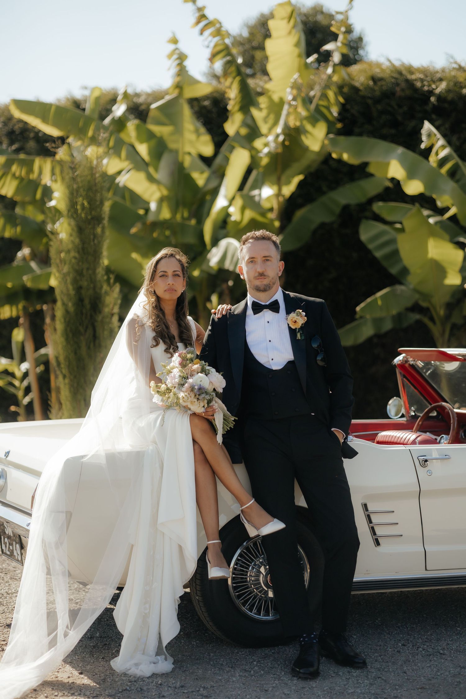 Elegant newlyweds pose with vintage white convertible car near tropical palm trees on their wedding day.
