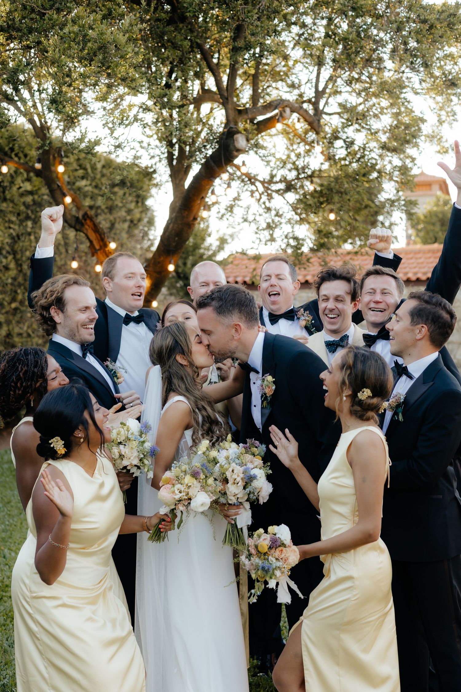 Wedding party celebrates outdoors with bridesmaids in yellow dresses and groomsmen in black tuxedos surrounded by trees.