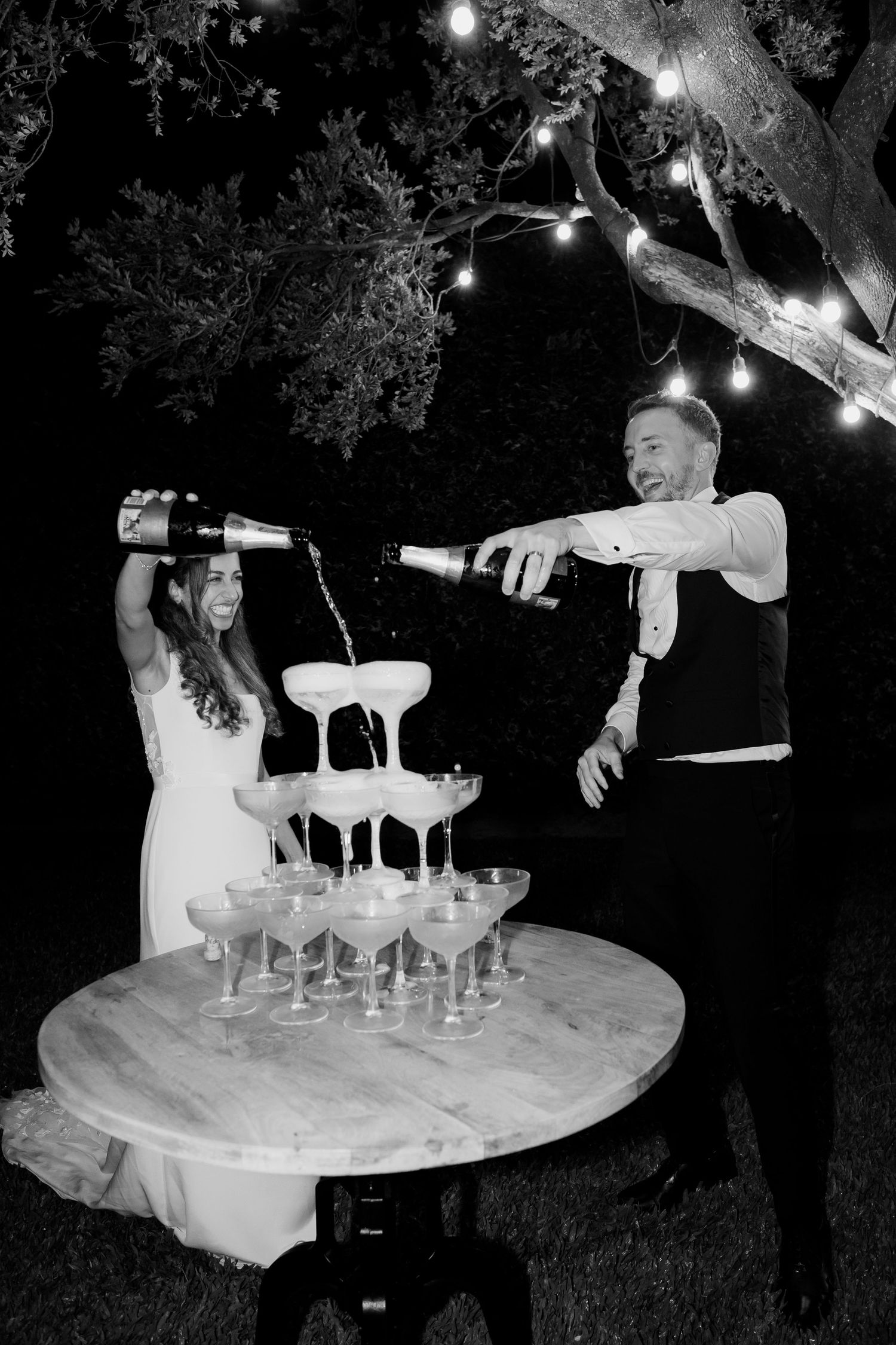 A couple pours champagne into a tower of glasses at an elegant outdoor nighttime celebration.