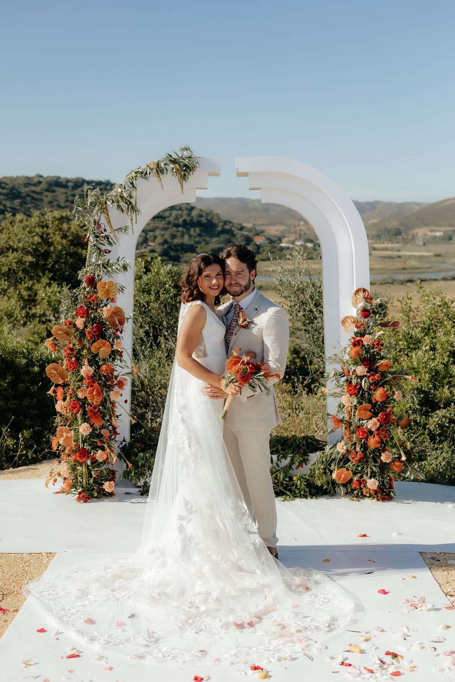 A wedding ceremony setup with a white arch decorated with orange flowers and greenery against a scenic countryside view.