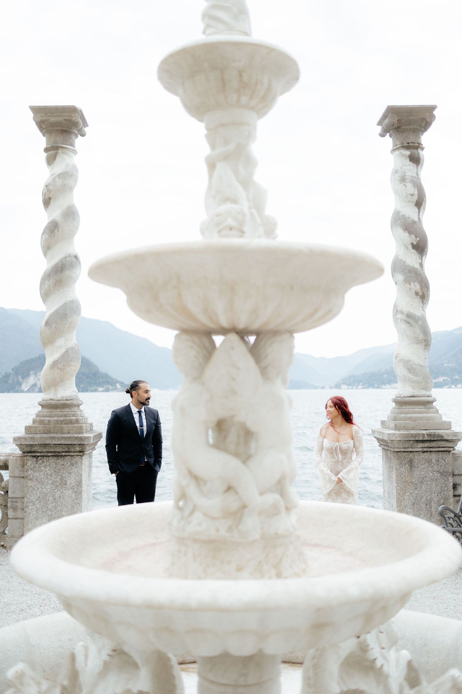 A romantic wedding scene with a grand white marble fountain and scenic mountain lake view in the background.
