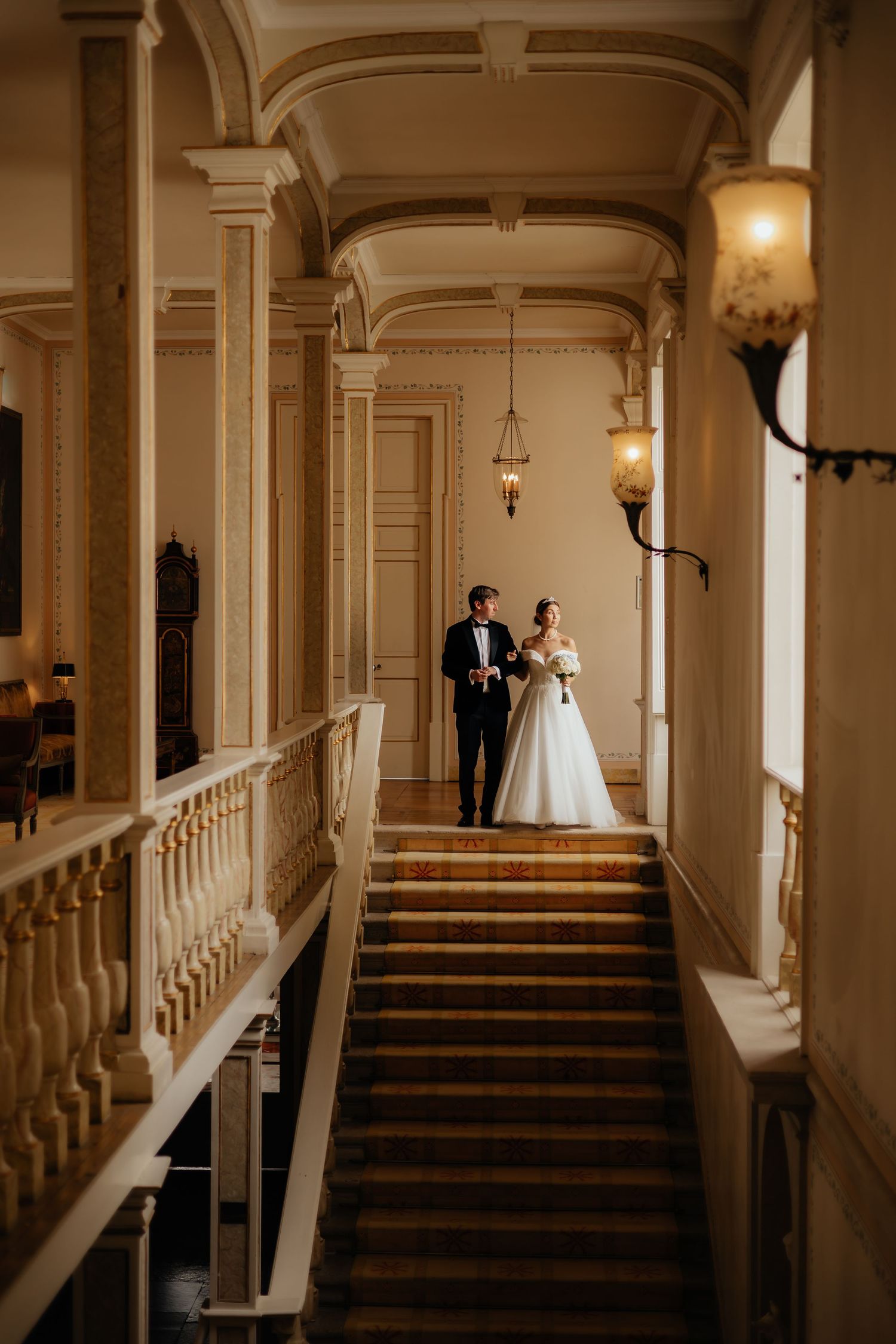 Elegant couple standing on grand staircase with ornate architectural details and vintage lighting.