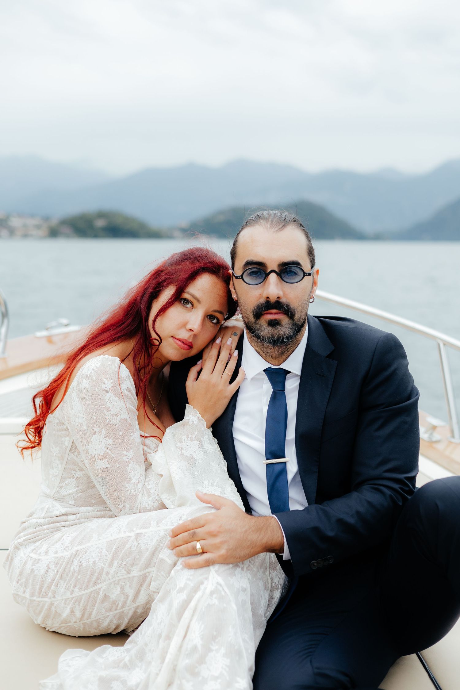 A stylish couple in wedding attire sitting on a luxury boat with mountains in the background.