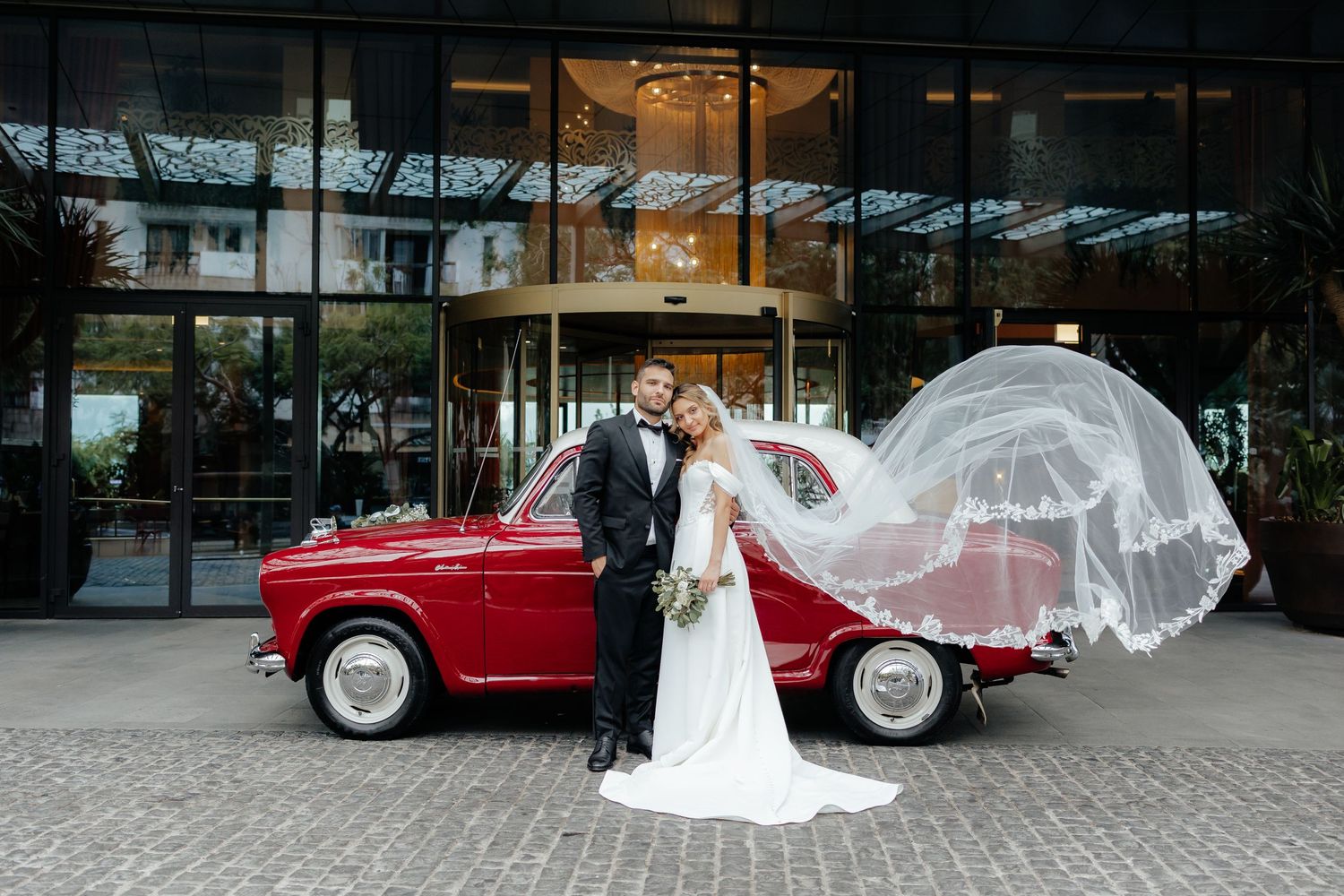 Newlyweds pose with a classic red car as the bride's veil dramatically flows in the wind outside a modern building.