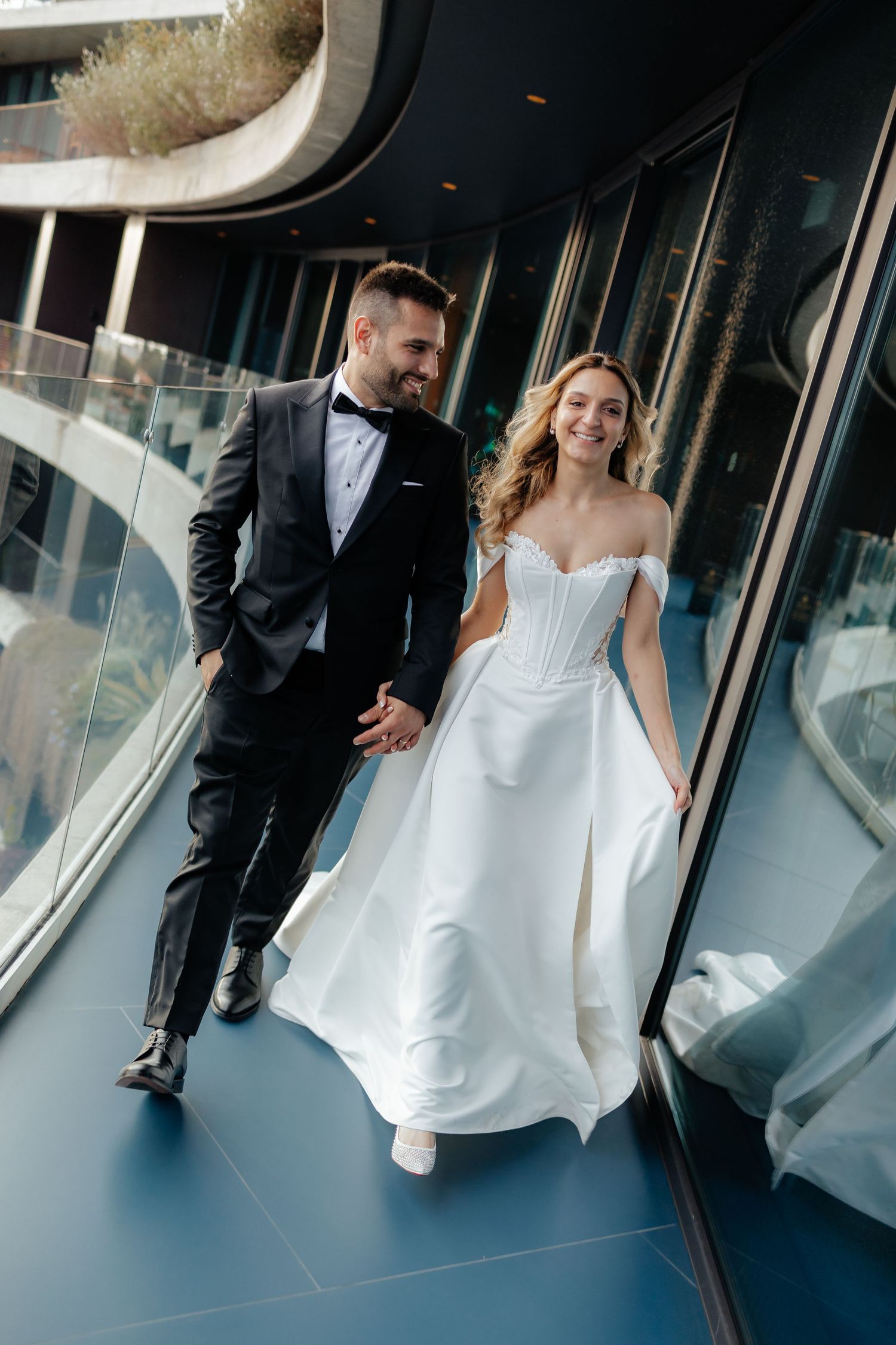 A couple walks hand in hand through a modern hallway, the bride's white dress flowing elegantly behind them.