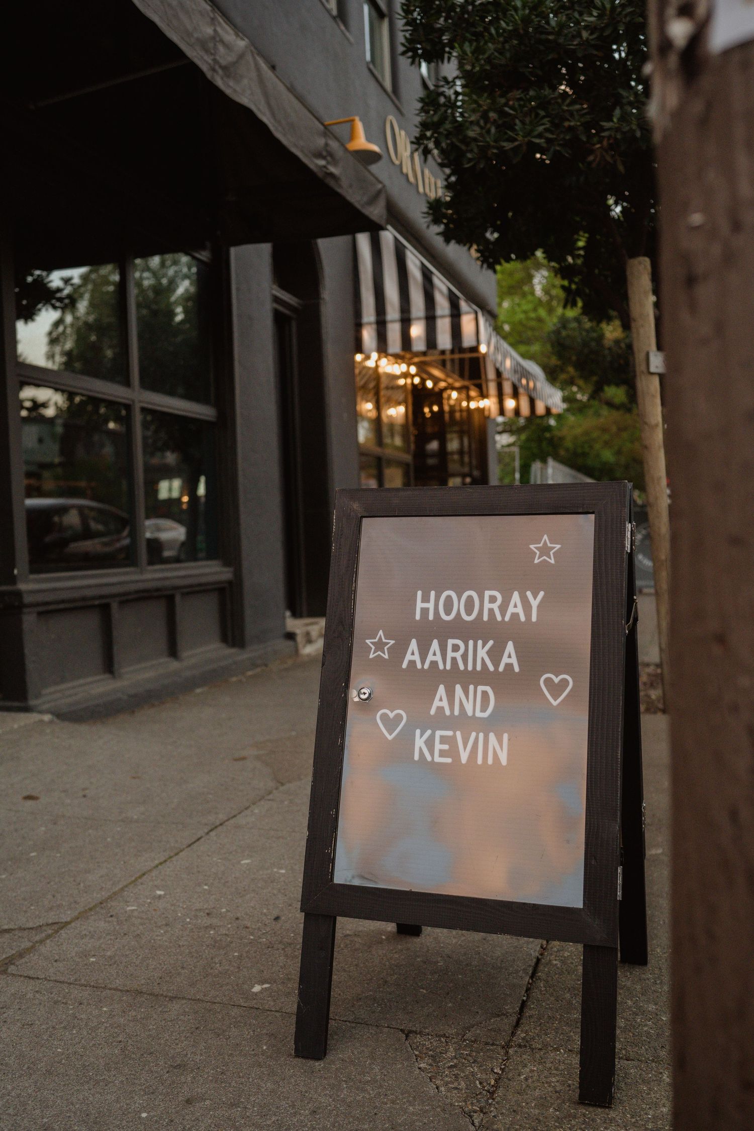 A chalkboard sign reading 'HOORAY AARIKA AND KEVIN' stands outside a venue with string lights.