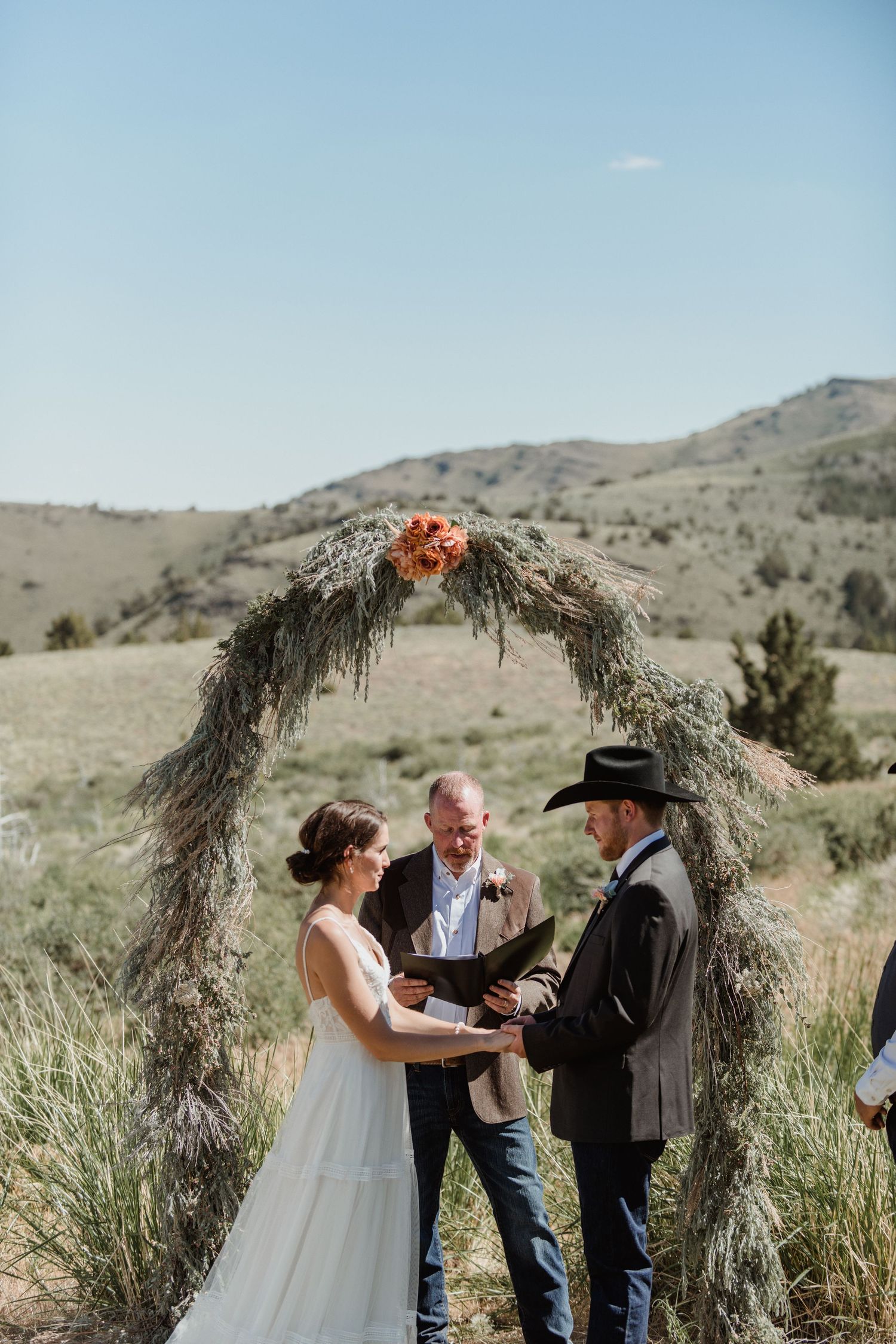 A rustic outdoor wedding ceremony takes place under a natural arch made of branches in a mountainous landscape.