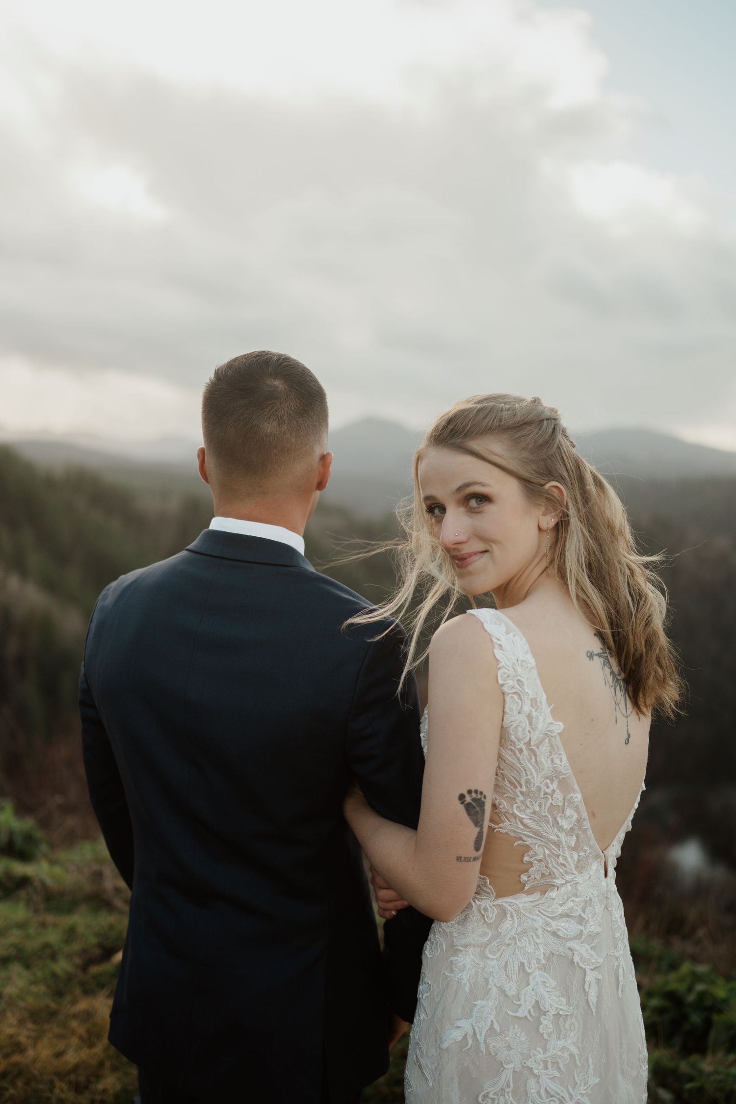 A couple in wedding attire stands on a hilltop overlooking mountain views.