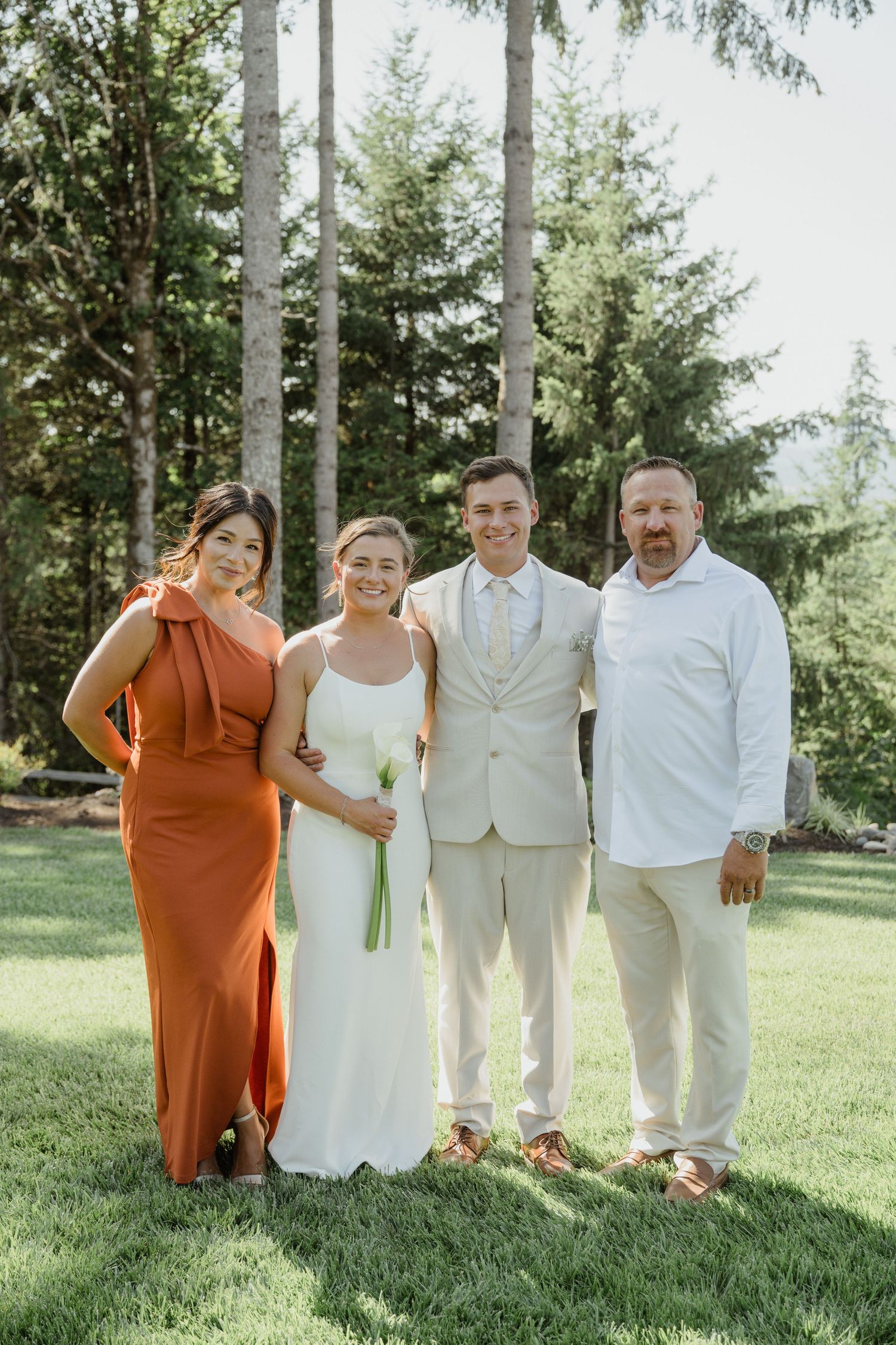 Four people pose together in formal attire against a backdrop of tall evergreen trees.